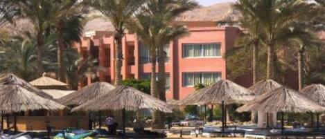 Un resort en la playa con palmeras, sombrillas de paja y un edificio con varios balcones.