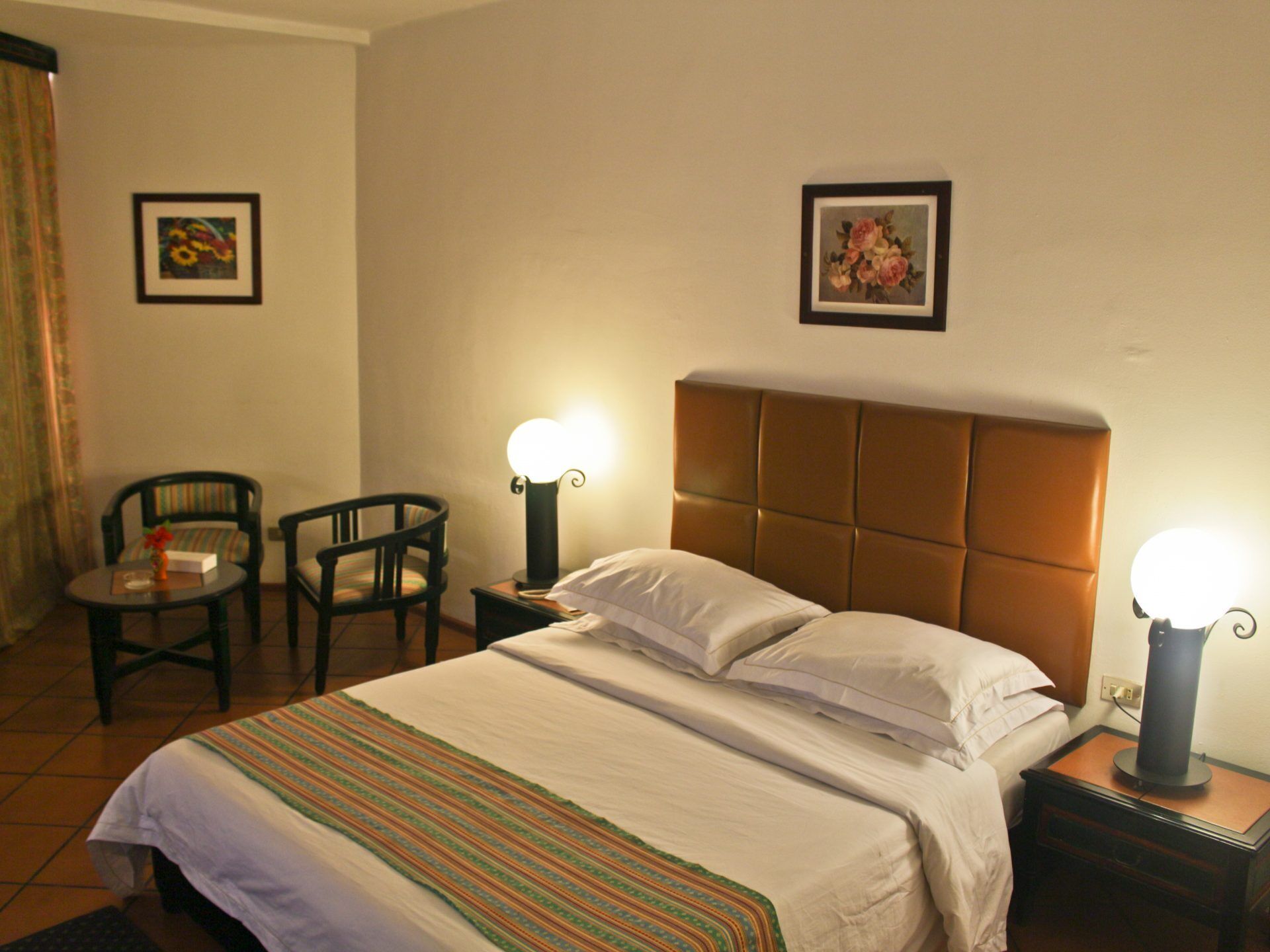 A hotel room with a bed, two chairs, a nightstand with a lamp, and two framed pictures on the wall.