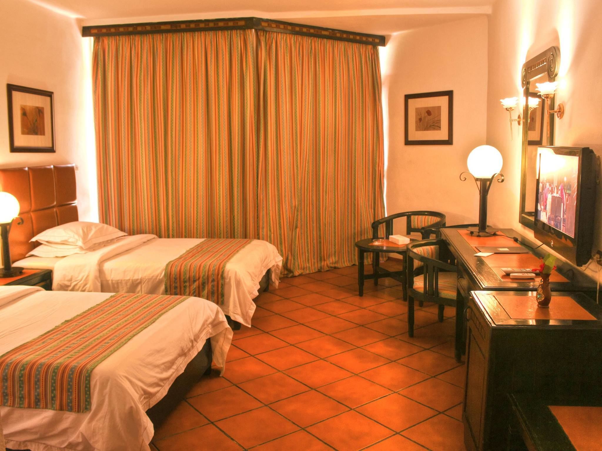 A hotel room with two beds, a desk, a chair, a TV, and a large window with curtains.
