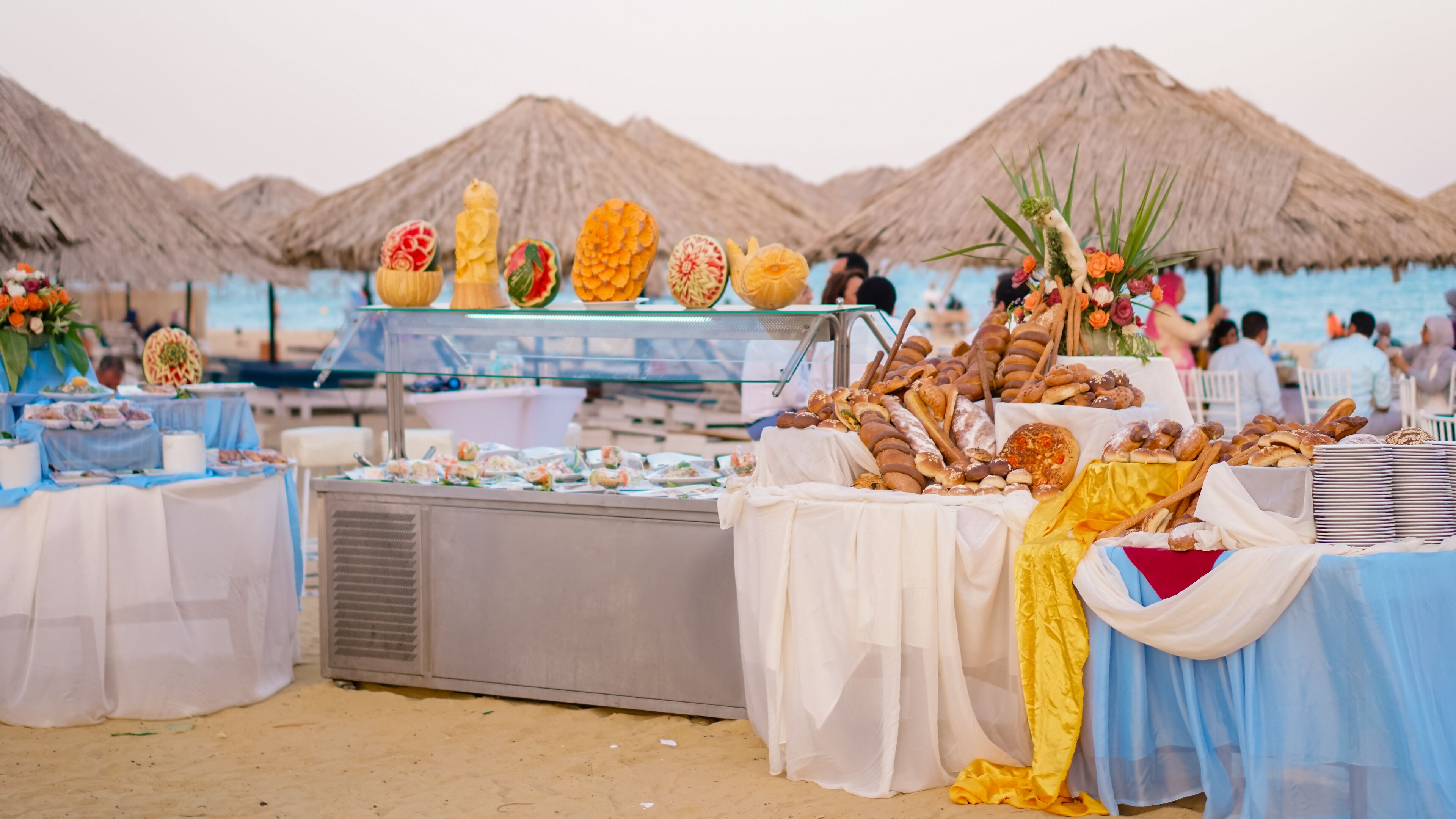 Un buffet proposant une variété de plats, agrémenté d’un centre de table décoratif, installé sur une plage où l’on aperçoit, au loin, des parasols en chaume.