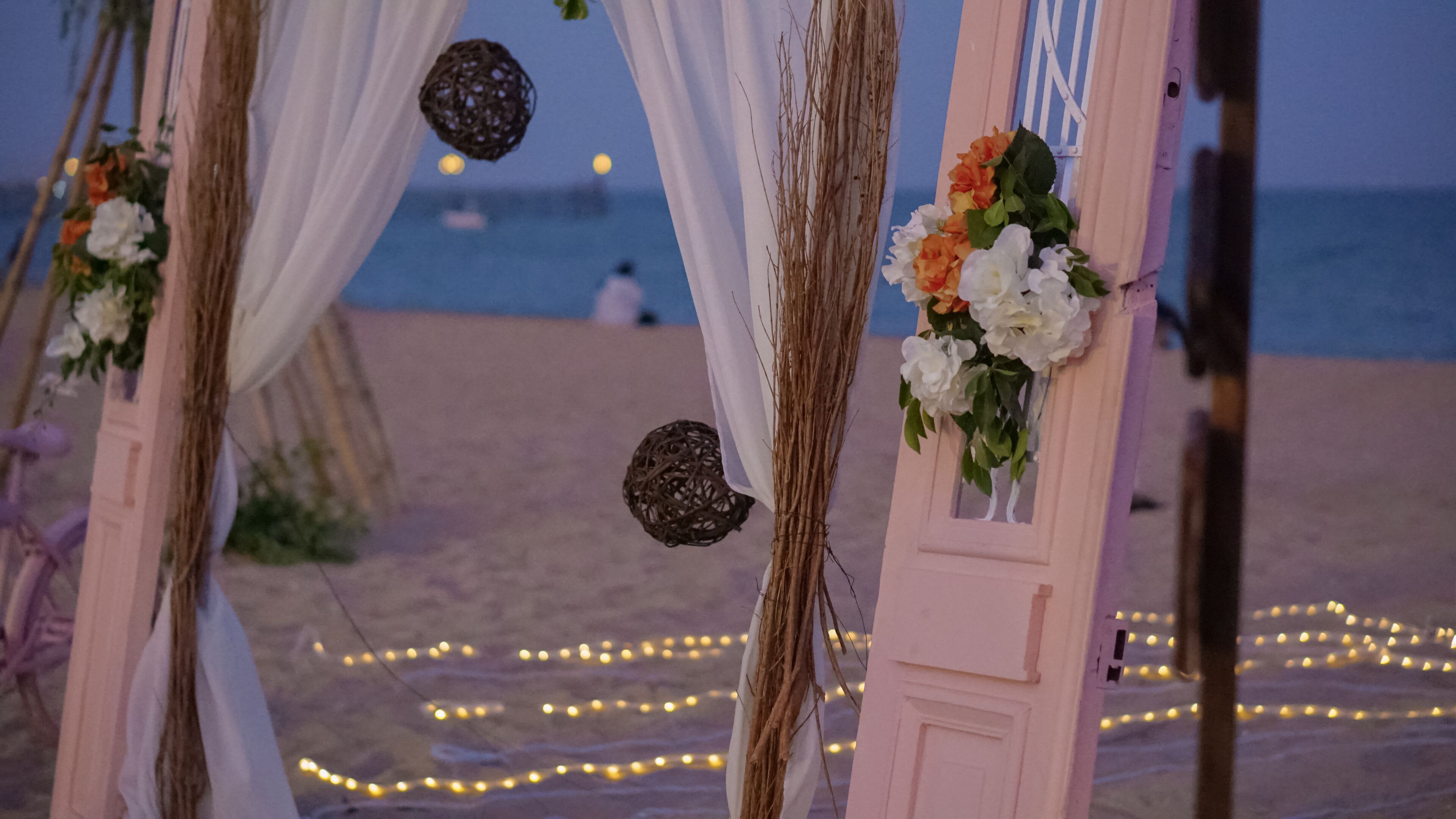 Een bruiloftsthrone op het strand met witte draperieën en bloemdecoraties, lichtjes en uitzicht op de oceaan.