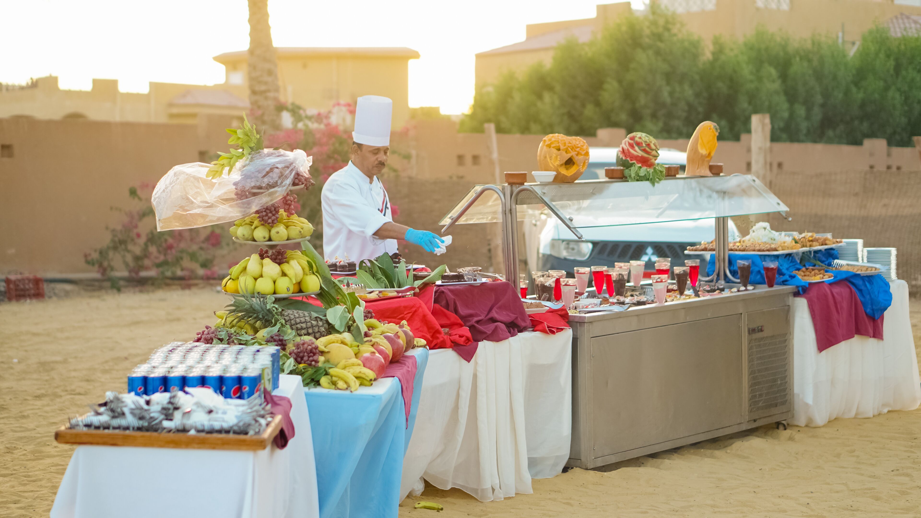 A food stall with a variety of fruits and desserts, set up on a sandy surface with buildings in the background.
