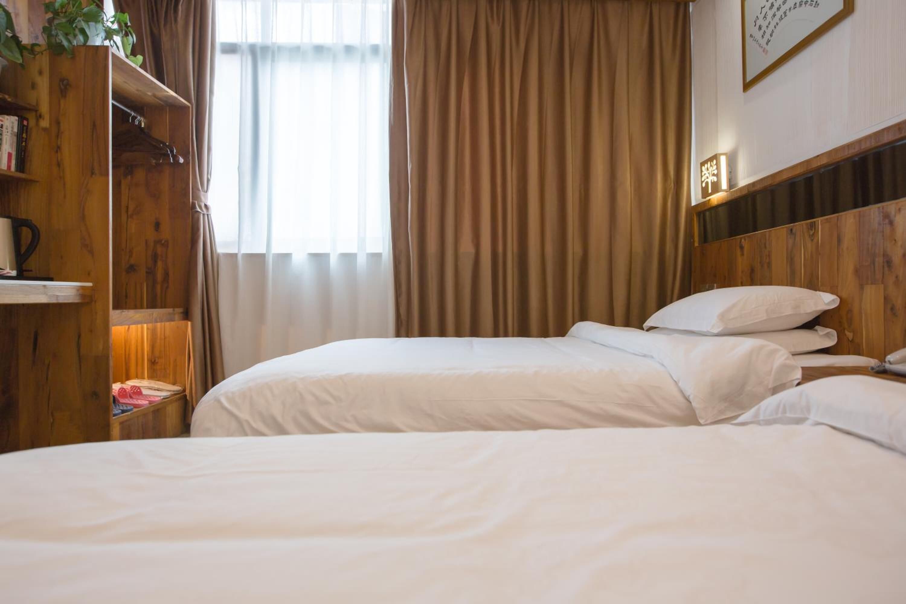 A hotel room with two beds, wooden headboard, a bookshelf, and a large window with curtains.