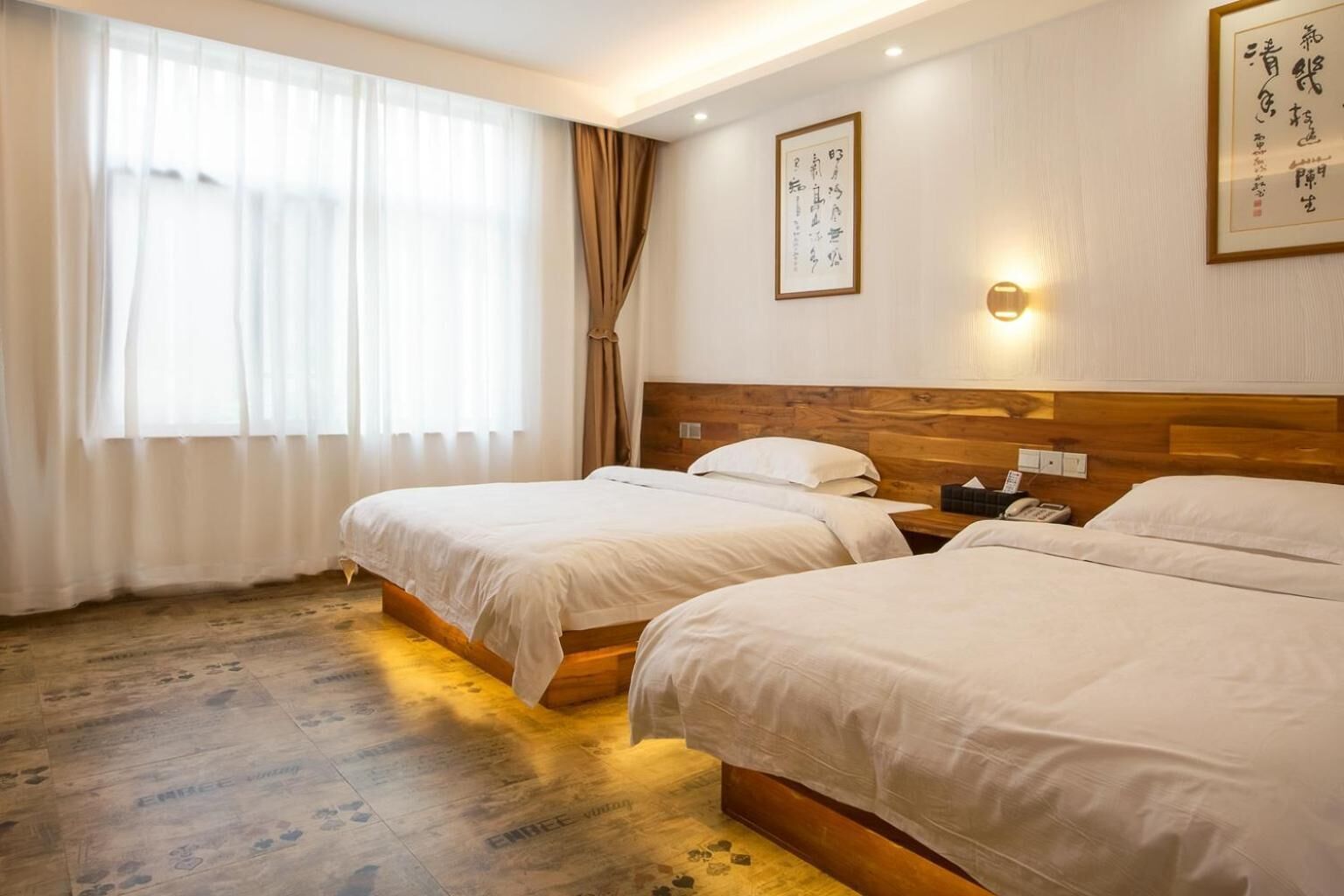 Ein Hotelzimmer mit zwei Betten, einem hölzernen Kopfteil, einem Nachttisch mit Telefon und einem Fenster mit durchsichtigen Vorhängen.