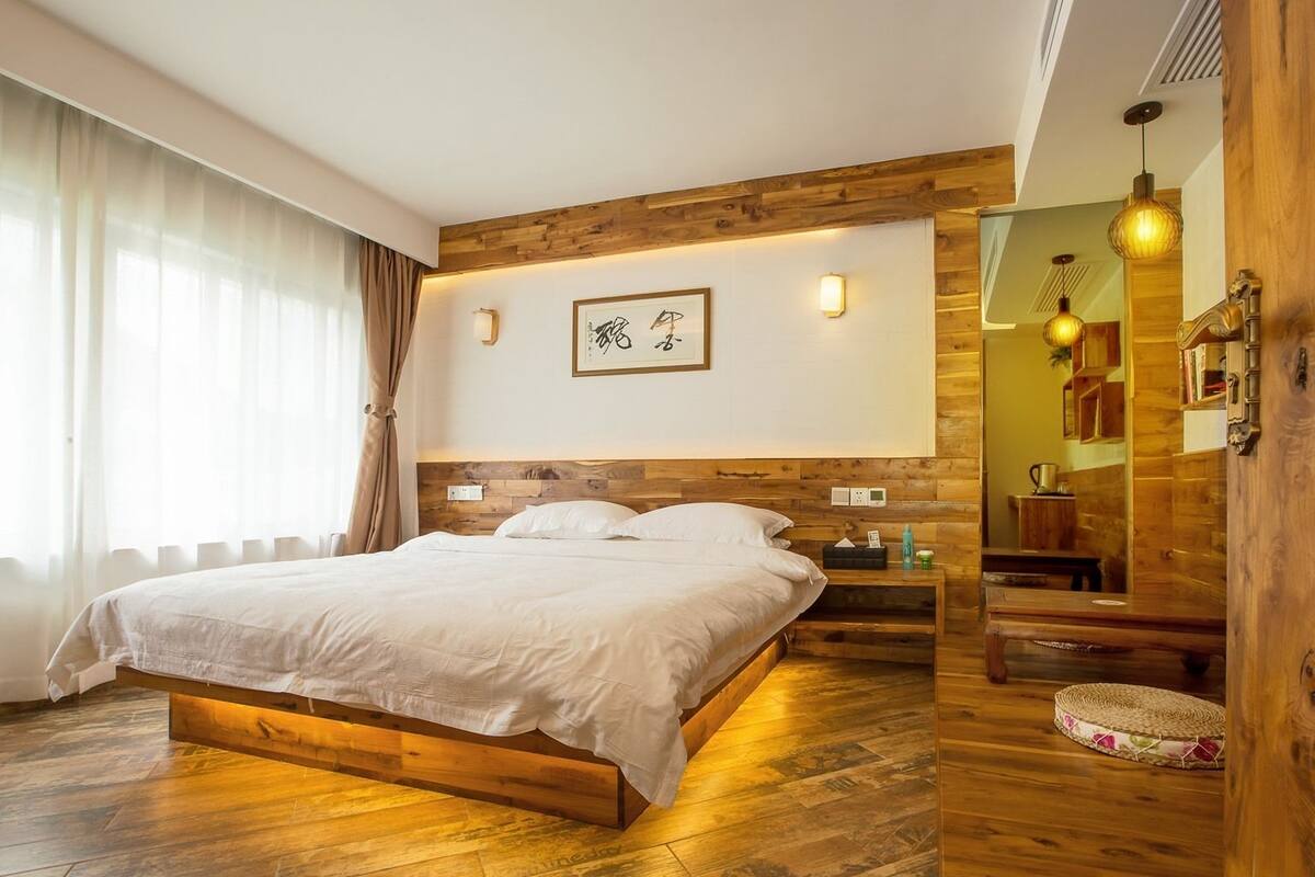 A bedroom with a wooden bed frame, a bedside table, a nightstand, a mirror, and a wall-mounted light fixture.