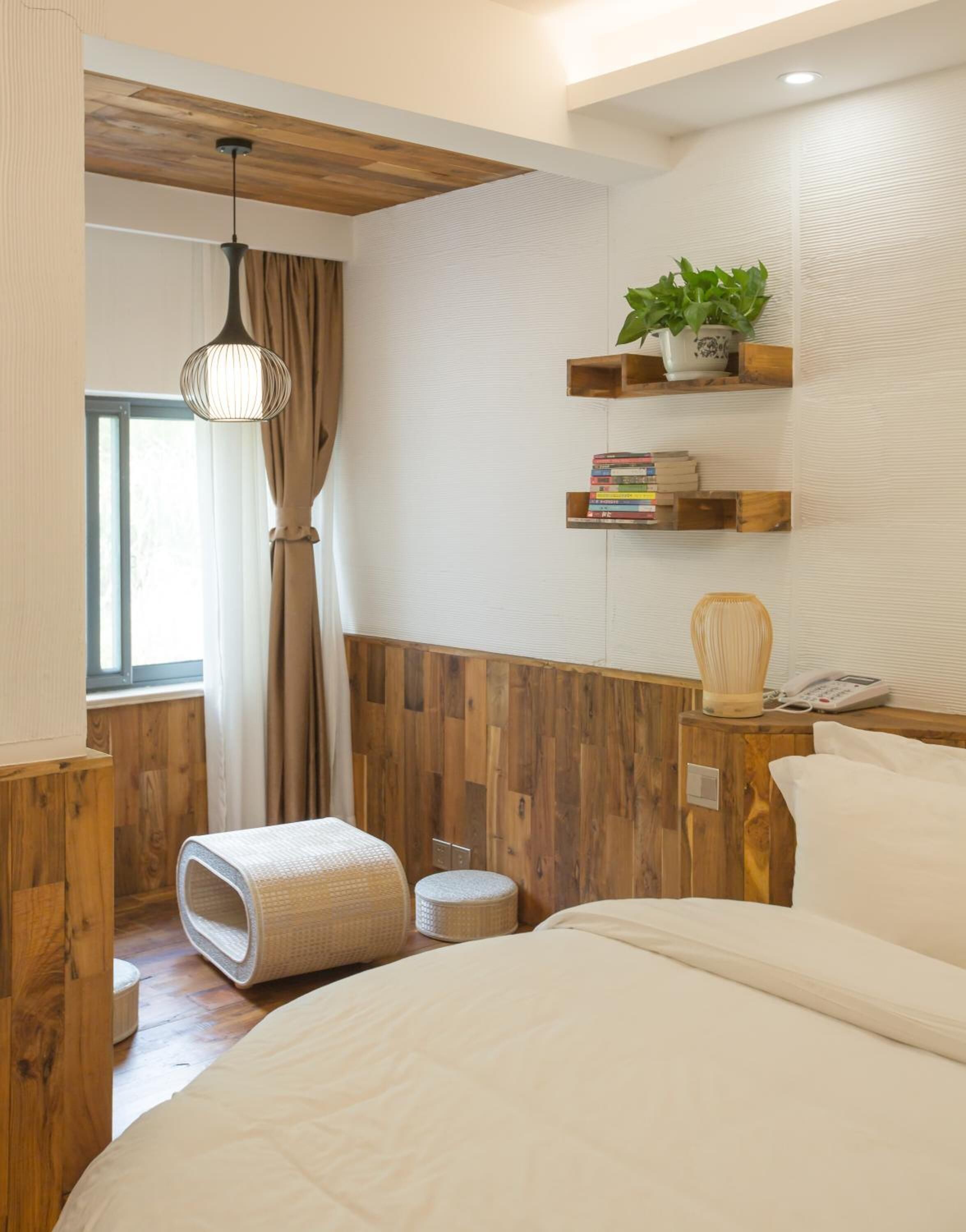 A bedroom with a wooden floor, a bed, a bedside table with a lamp, a shelf with books and a plant, and a window with curtains.