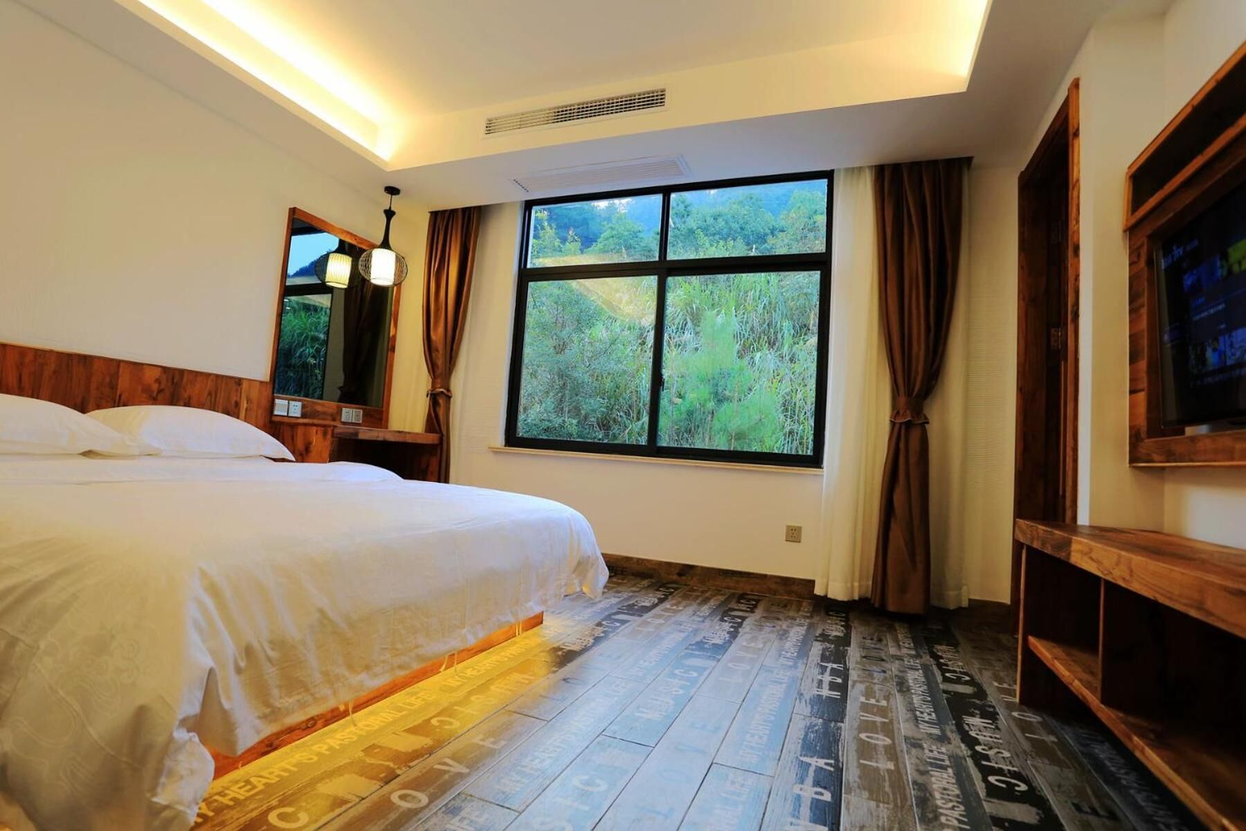 Un dormitorio con una cama grande, una ventana con vistas a la vegetación, un cabecero de madera y un televisor en la pared.
