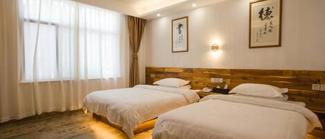 A hotel room with two single beds, a wooden headboard, a large window with sheer curtains, and two framed calligraphy pieces on the wall.