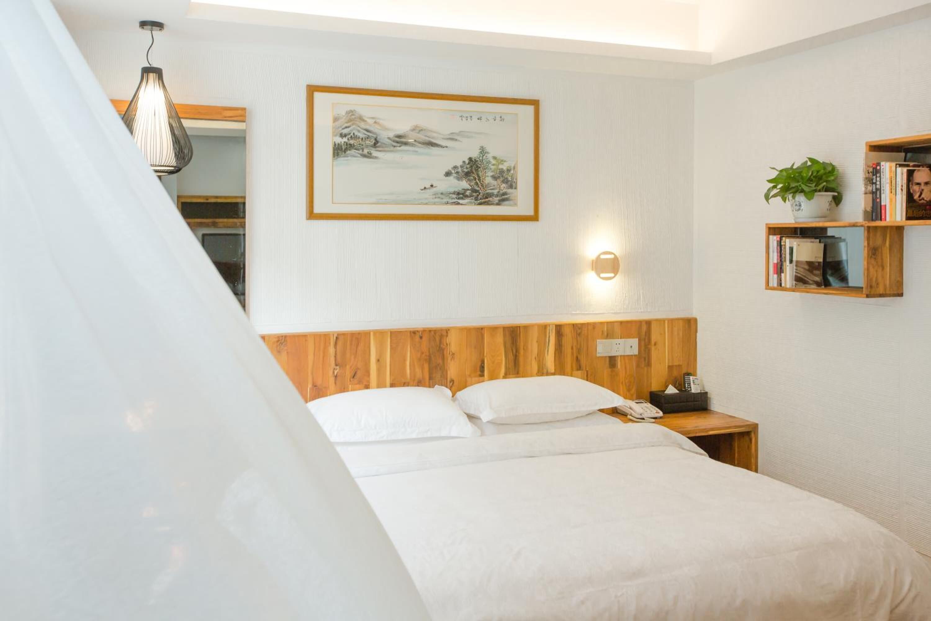Un dormitorio ordenado con cabecera de madera, una mesita de noche con lámpara, un cuadro enmarco y una pequeña estantería con libros y una planta.