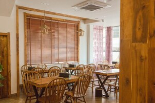 A dining area with wooden tables and chairs, large windows with blinds, and a ceiling vent.