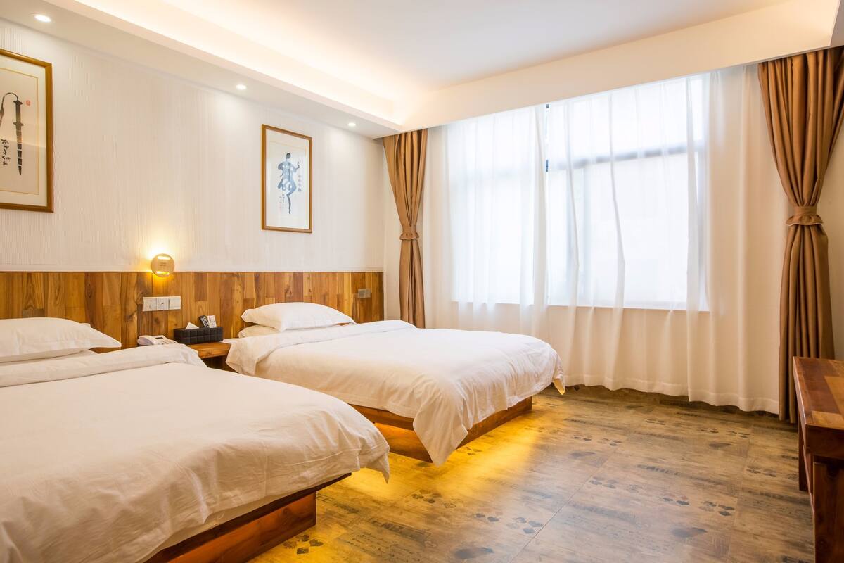 A hotel room with two beds, wooden flooring, a nightstand with a lamp, and a large window with curtains.