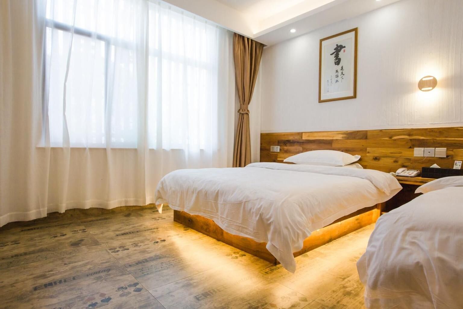 A hotel room with two beds, a wooden headboard, a bedside table with a phone, and a window with sheer curtains.