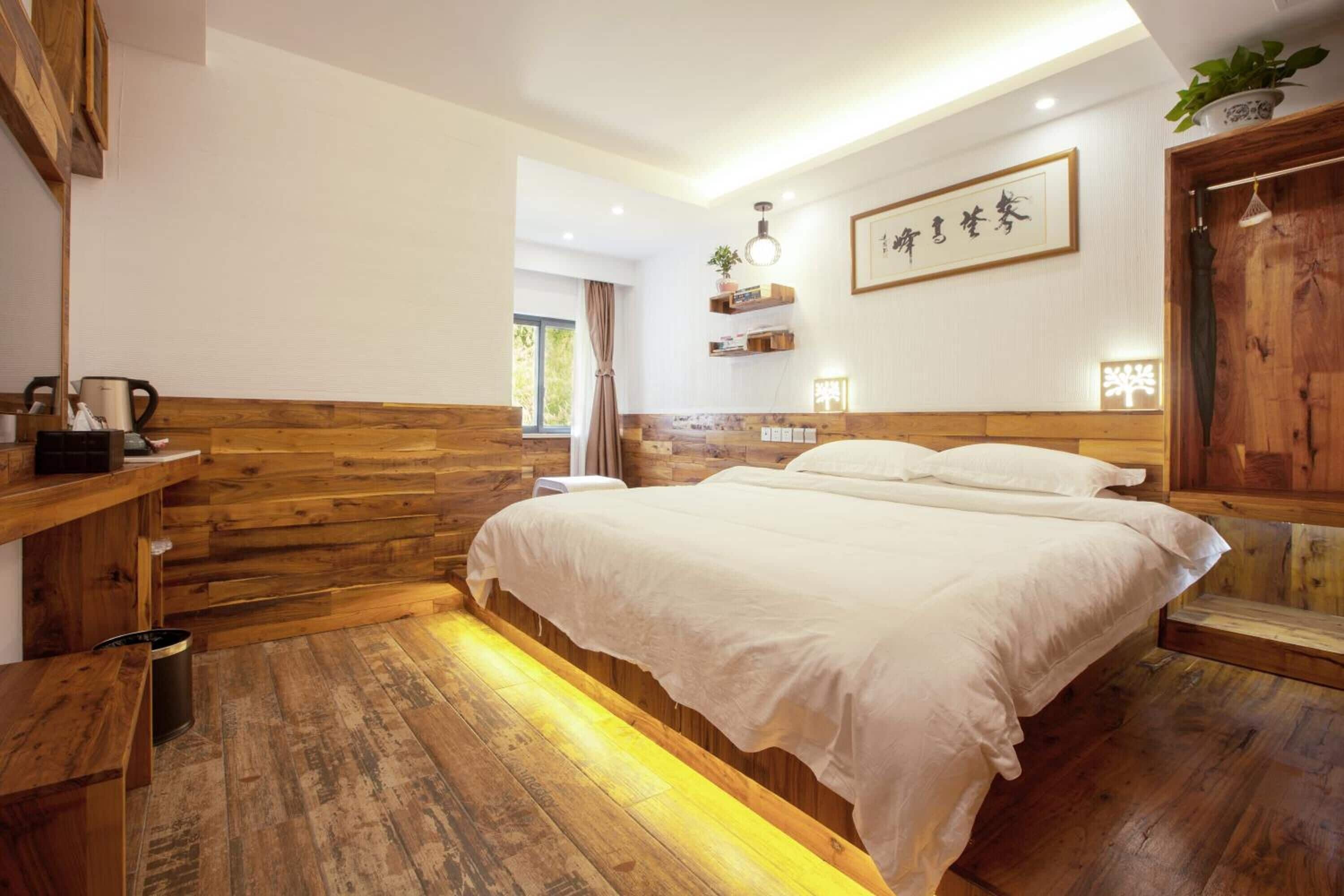 A bedroom with a wooden headboard, a bed with white bedding, a bedside table, a shelf with books and a plant, and a wall-mounted light fixture.