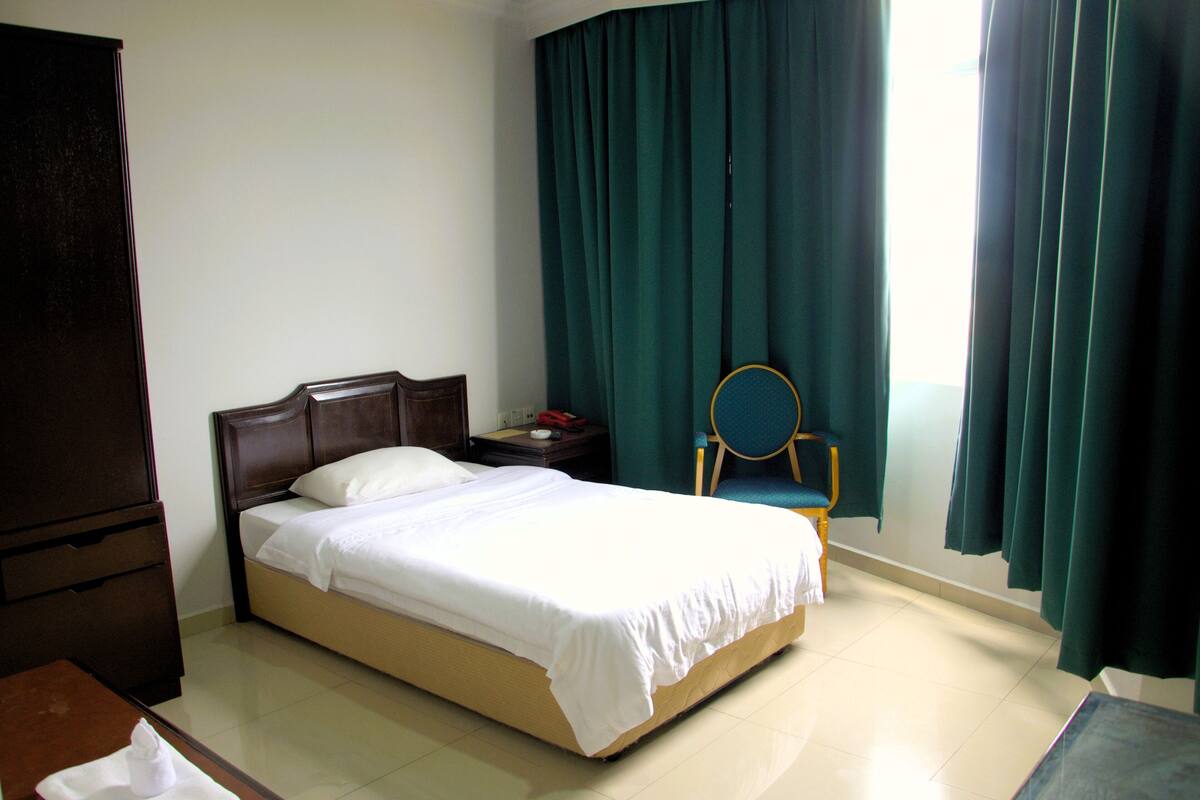 Un lit bien fait dans une chambre d’hôtel, avec une tête de lit en bois, une table de chevet avec une lampe et une chaise dotée d’un dossier arrondi.