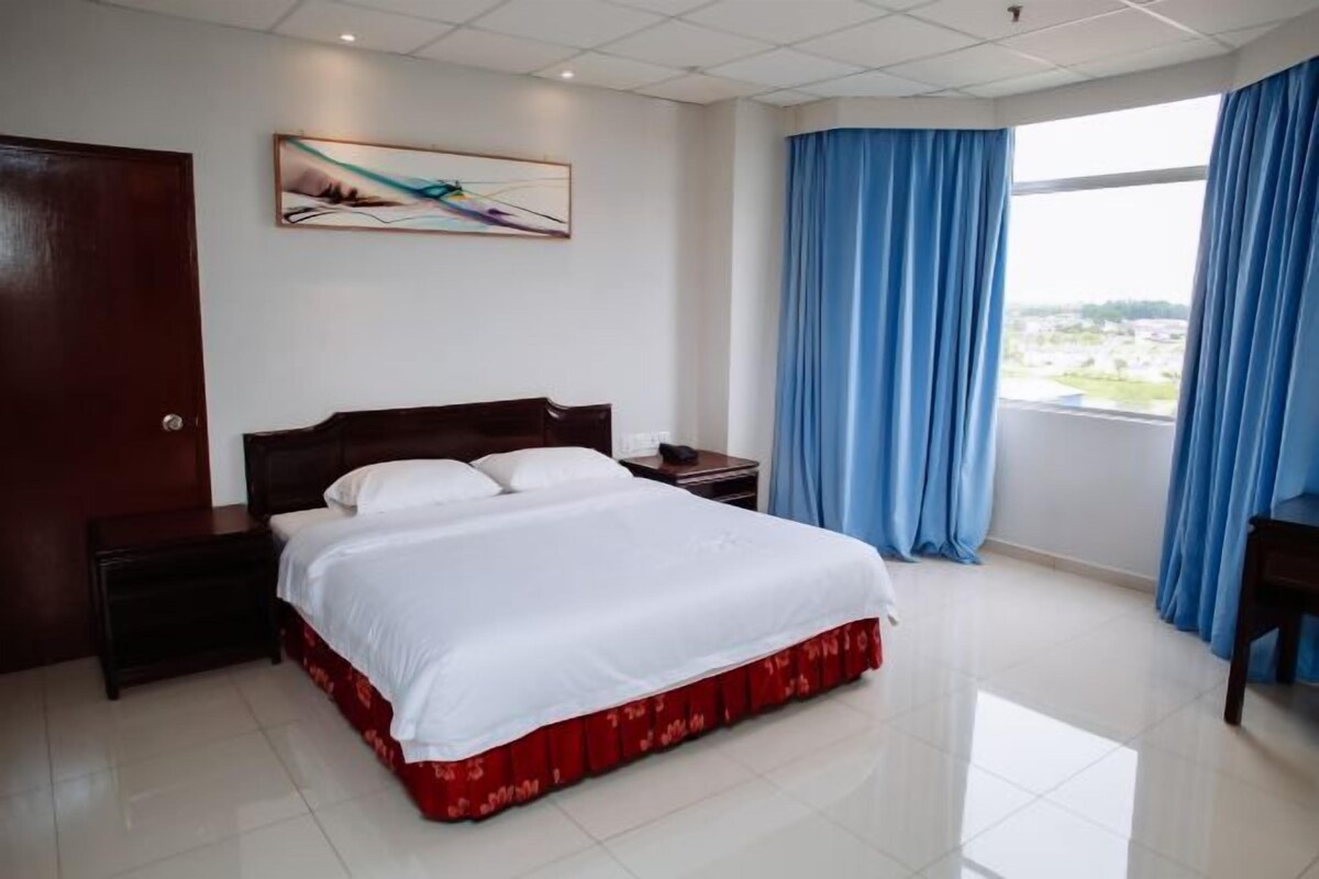 Een hotelkamer met een bed, nachtkastje, schilderij aan de muur en een raam met gordijnen.