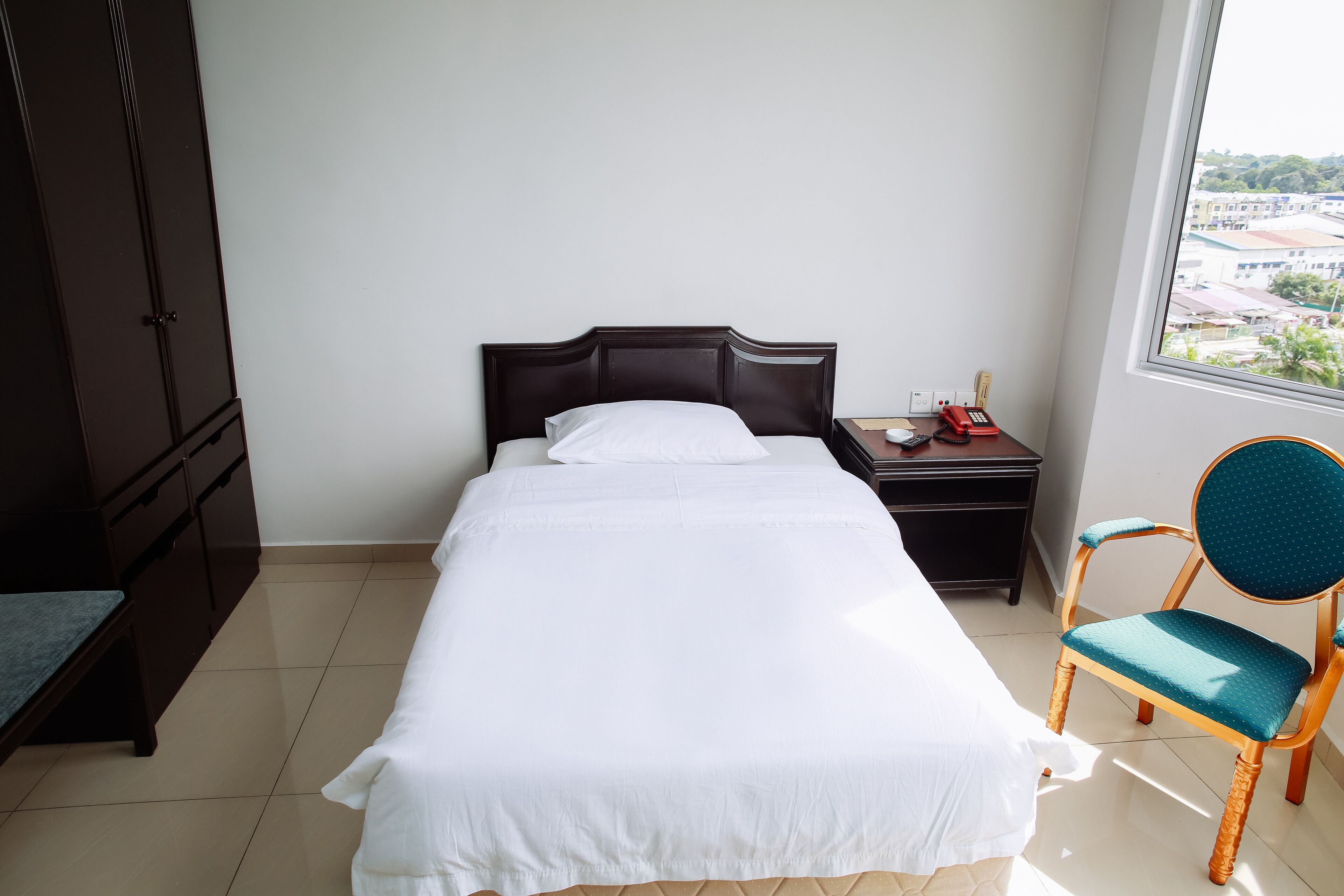A neatly made bed with white linens, a wooden nightstand with a red telephone, and a teal chair.