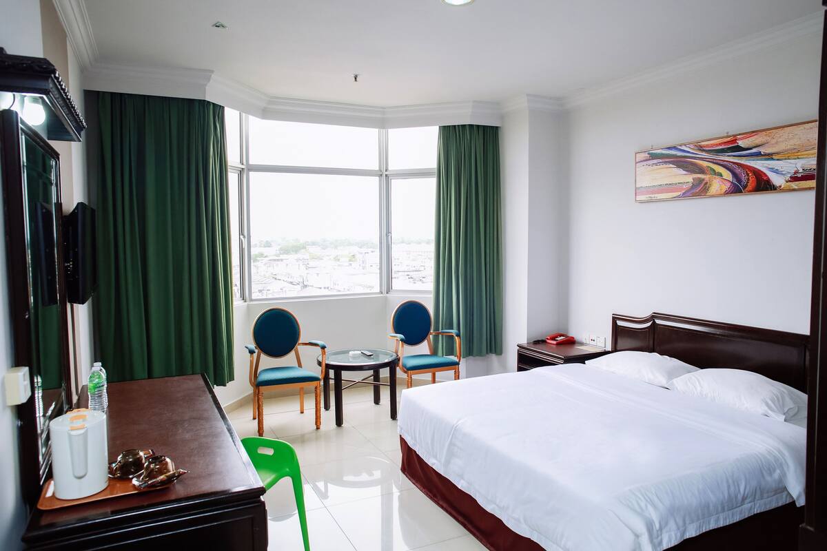 Une chambre d’hôtel comprenant un lit, un bureau avec une chaise, une petite table avec deux chaises et une grande fenêtre avec des rideaux.