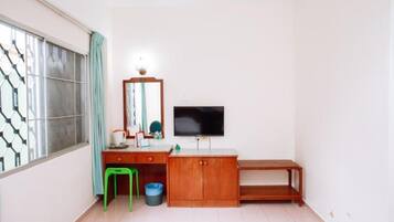 Una habitación pequeña con un escritorio de madera, un taburete verde, un televisor y una ventana con un diseño geométrico.