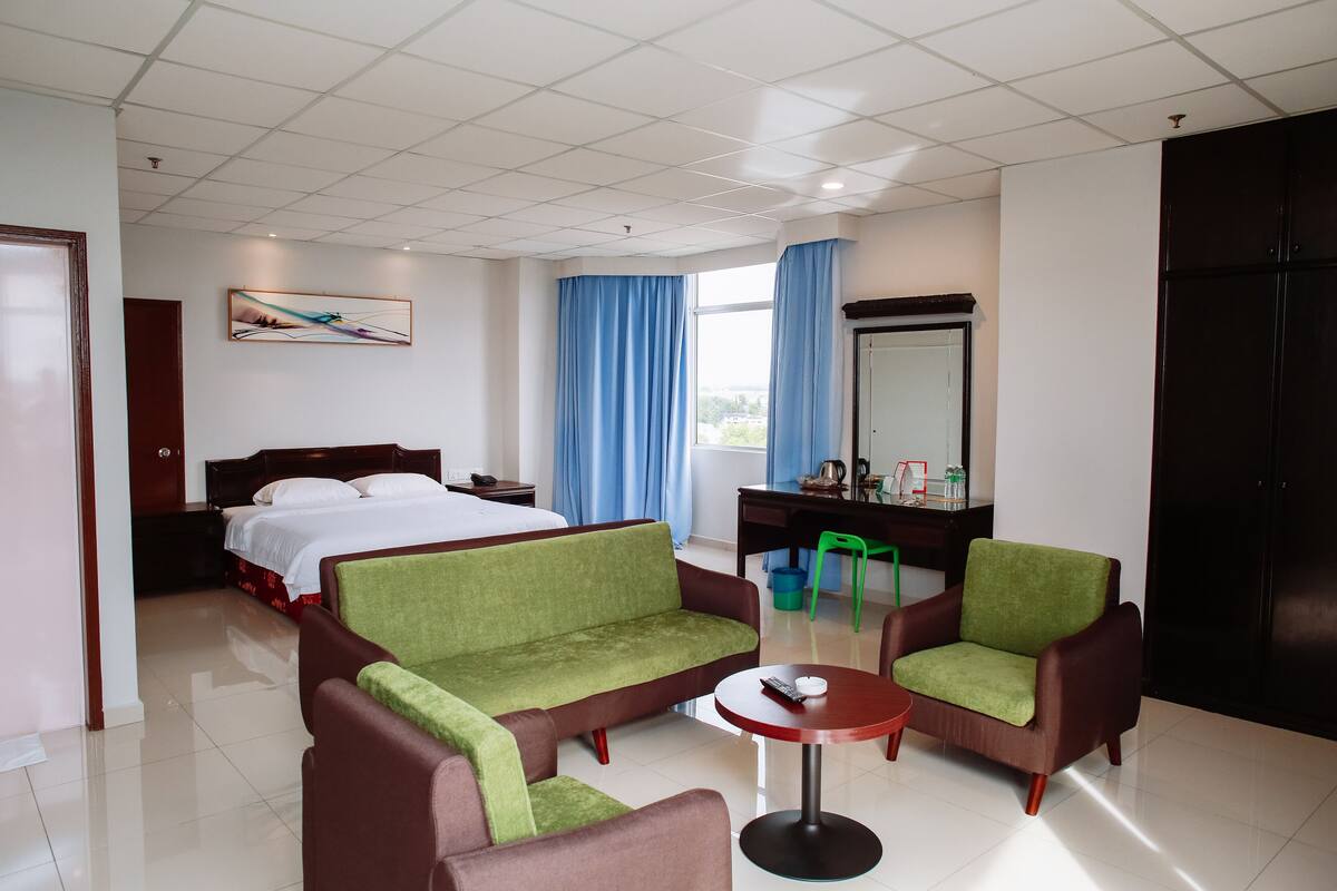 Une chambre d’hôtel avec un lit, un canapé vert, deux fauteuils, une petite table, un bureau, un miroir et une fenêtre avec des rideaux.