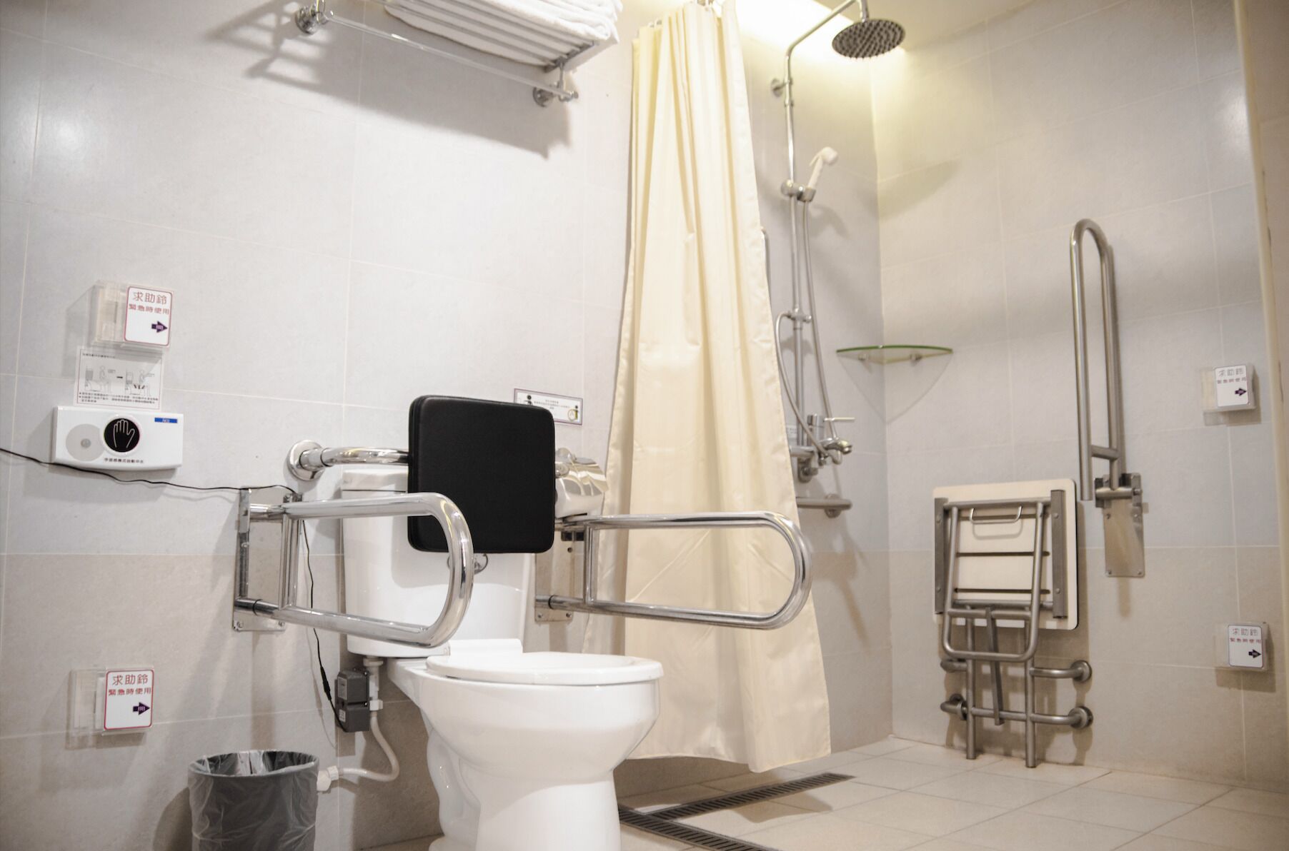 Une salle de bain équipée de toilettes, d’une douche, de barres d’appui et d’un distributeur de gel hydroalcoolique.
