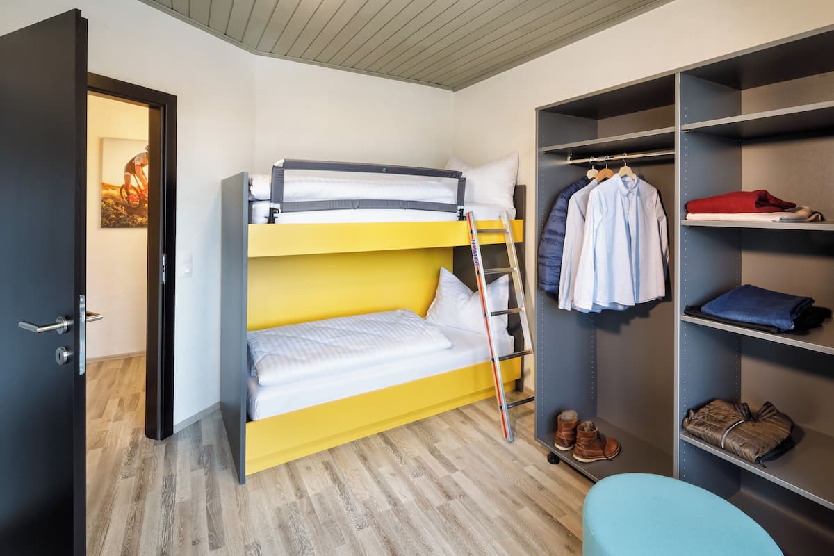 Ein Schlafraum mit Etagenbett, einem gelben Unterbett, einem Kleiderschrank mit darin hängender Kleidung und einem Paar Stiefeln auf dem Boden.