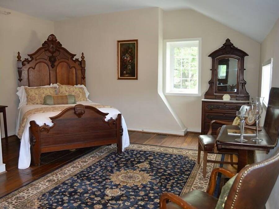 Une chambre à coucher avec un grand lit en bois, une commode classique, un miroir et une fenêtre laissant entrer la lumière naturelle.