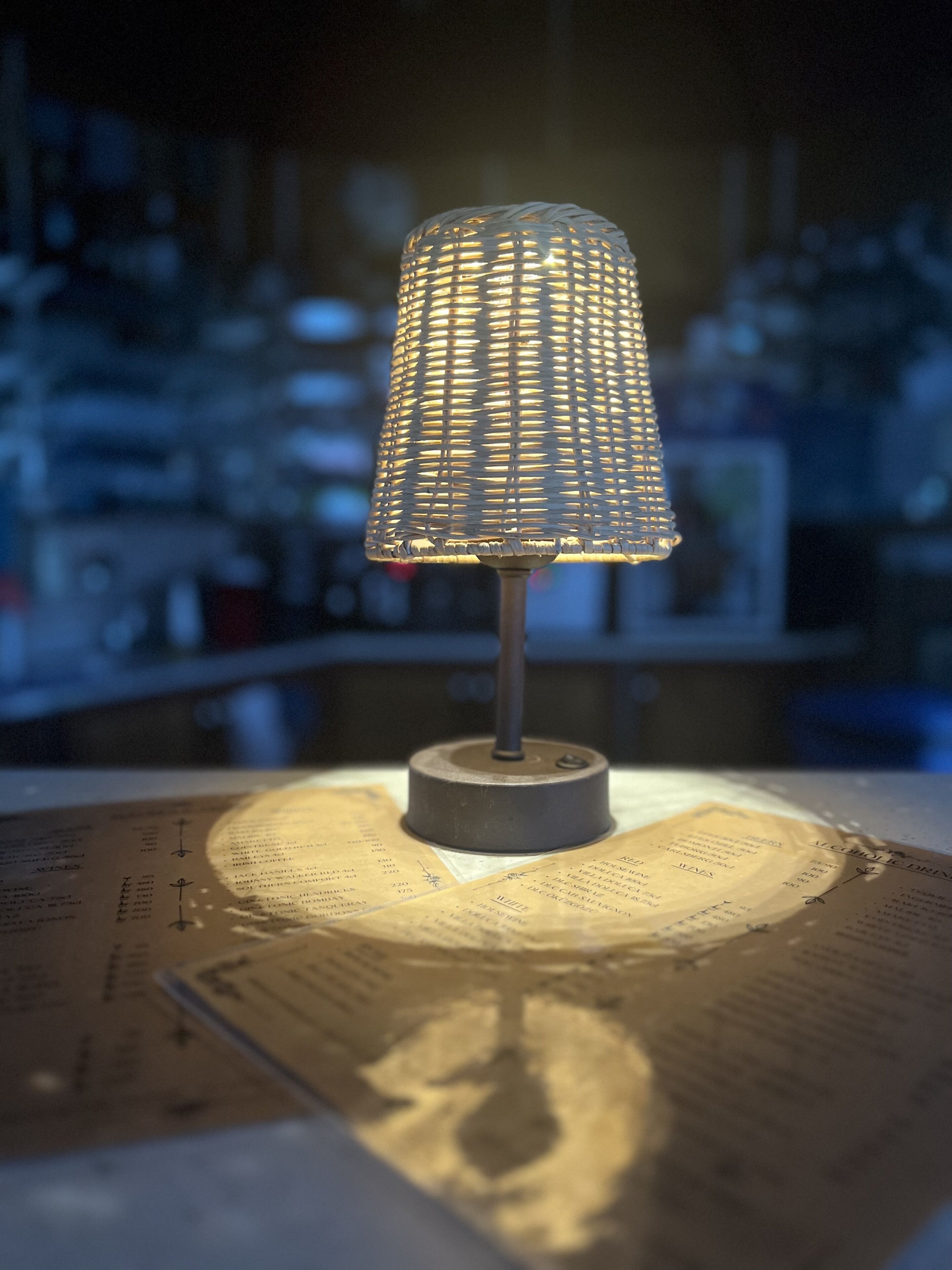 Eine Tischlampe mit gewebtem Schirm auf einem Tisch, unter dem eine Speisekarte liegt.