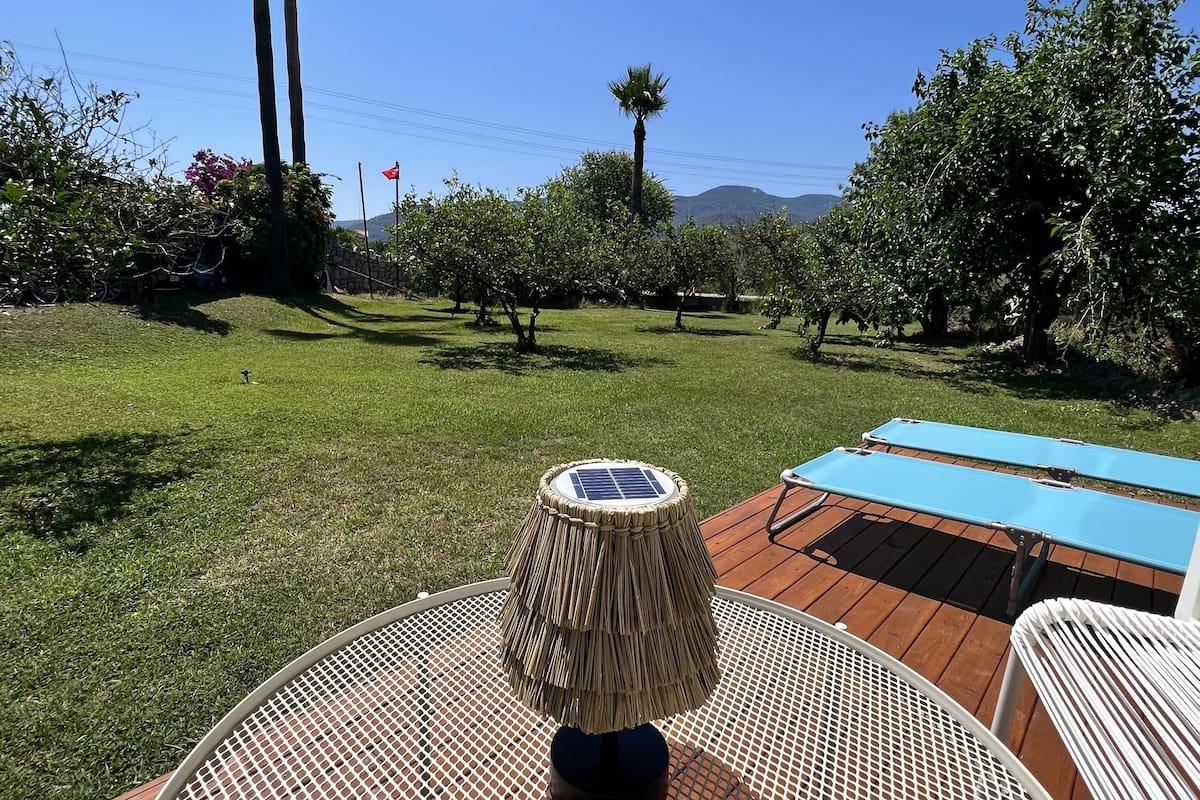 A round metal table with a woven shade, surrounded by a wooden deck, with a view of a grassy area and trees in the background.
