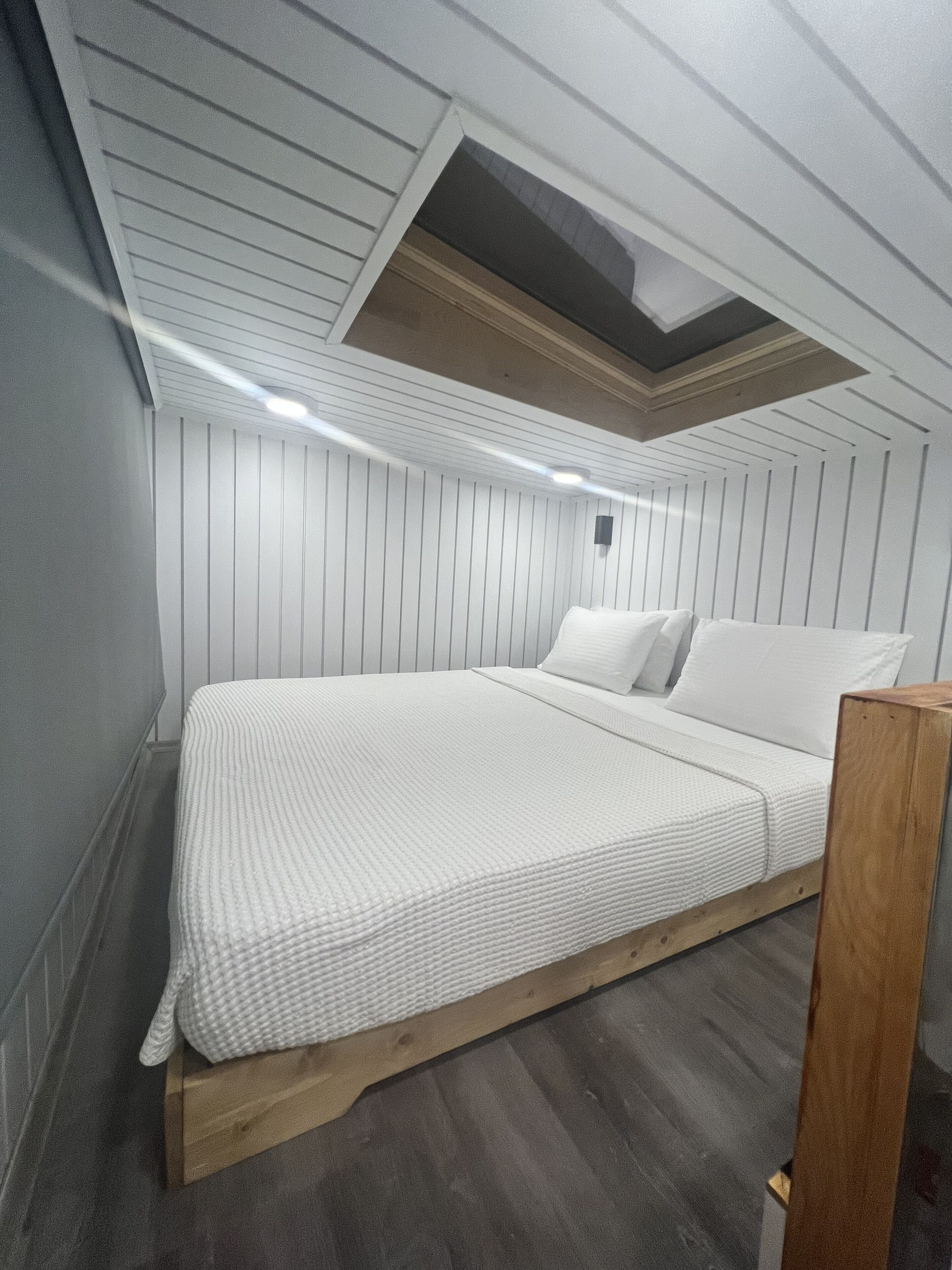A bedroom with a sloped ceiling, wooden bed frame, white bedding, and a skylight.