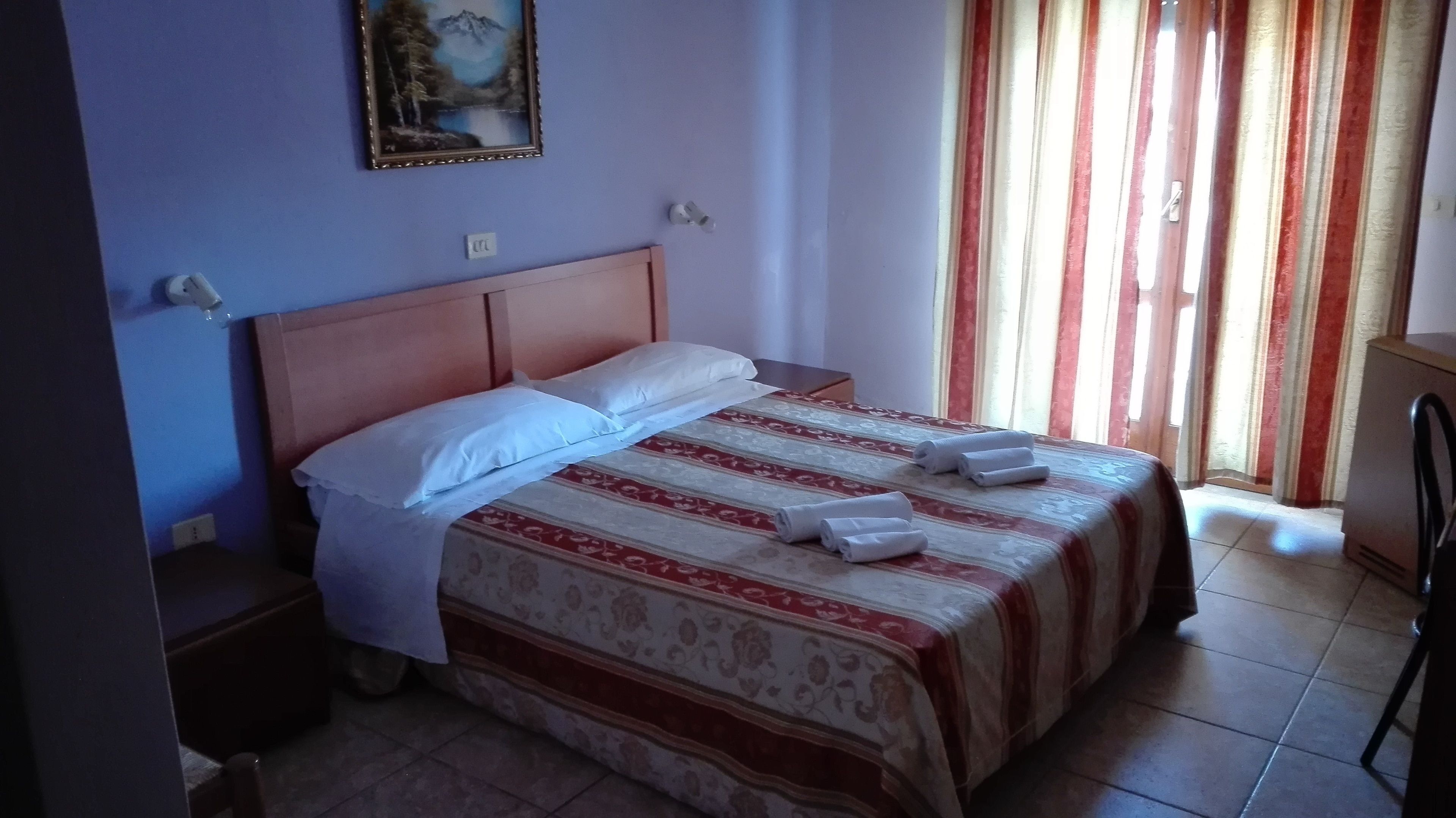 Habitació d'hotel amb llit, una tauleta de nit, un quadre a la paret i una finestra amb cortines.