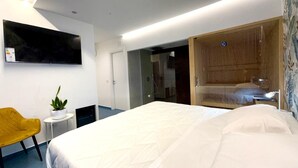 A modern hotel room with a large bed, a small side table with a plant, a yellow chair, a wooden wardrobe, and a TV mounted on the wall.