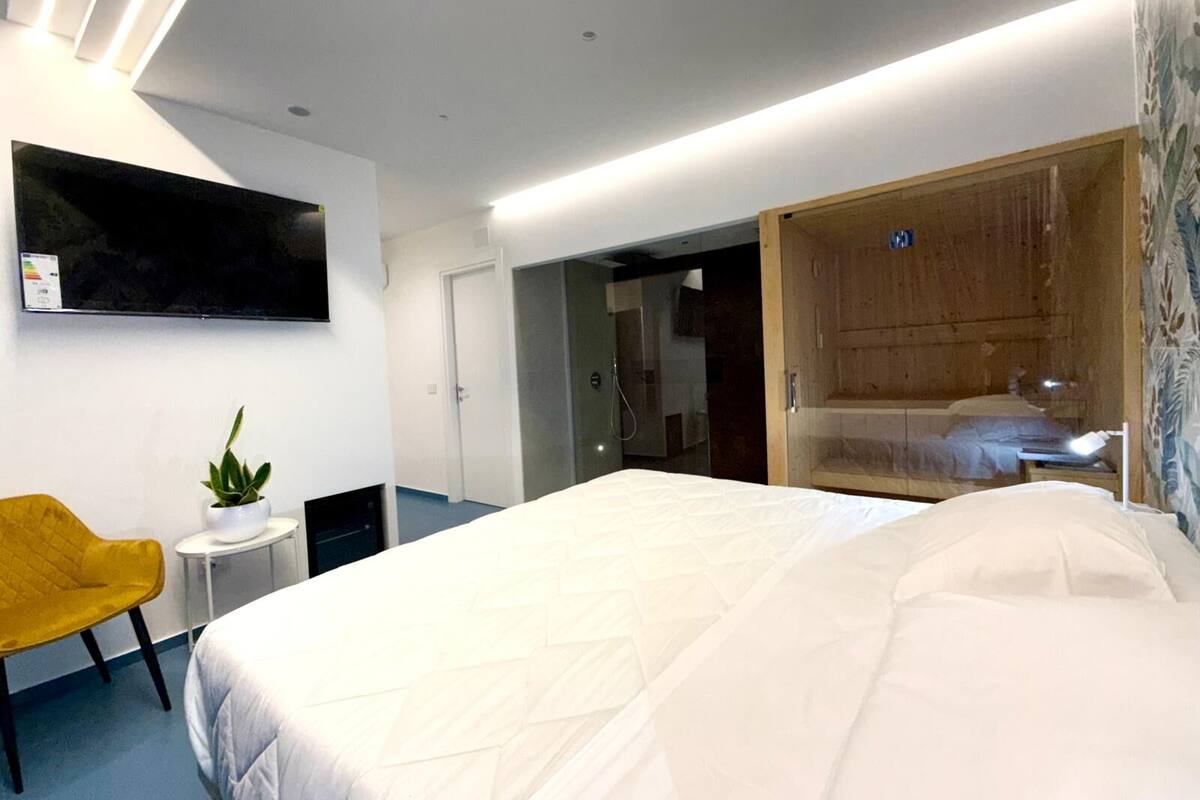 Ett modernt hotellrum med en stor säng, ett litet sidobord med en växt, en gul stol, en trägarderob och en tv som är monterad på väggen.