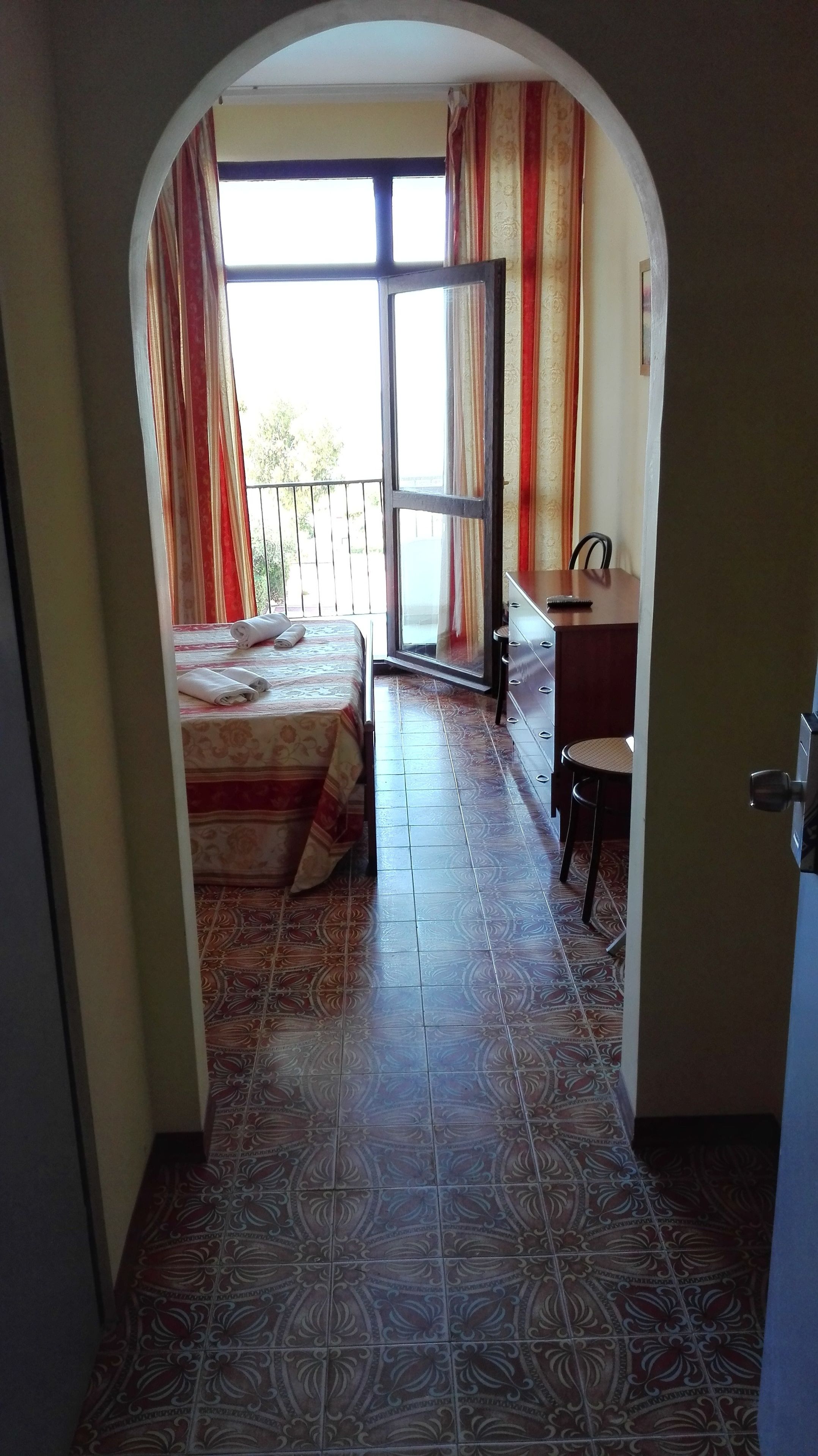 A room with a bed, a desk, and a chair, with a balcony visible through an open door.