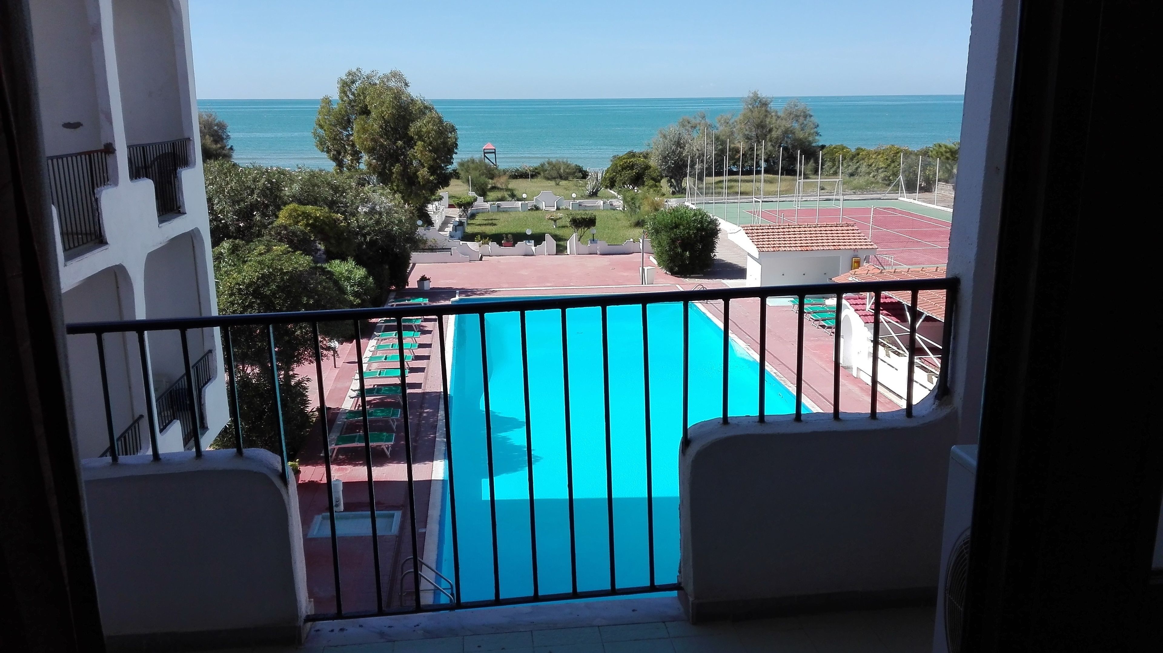 A balcony with a view of a swimming pool, a tennis court, and the sea.
