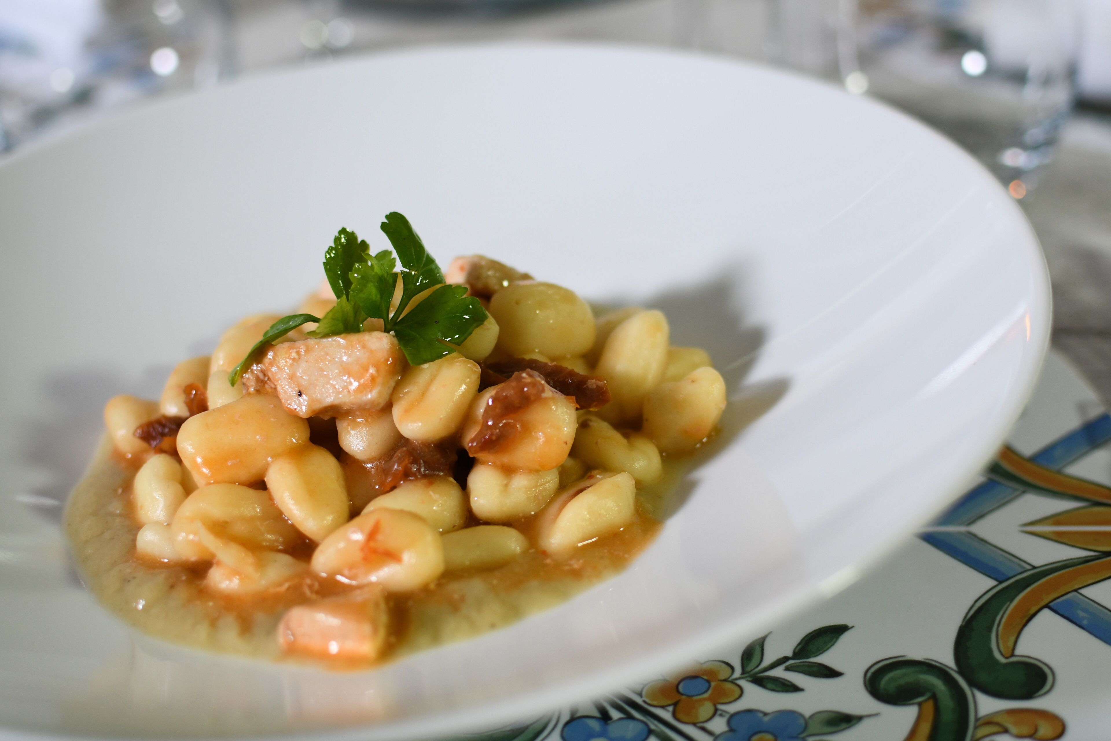 A white plate with a dish of beans and meat, garnished with parsley, on a table with a patterned tablecloth.