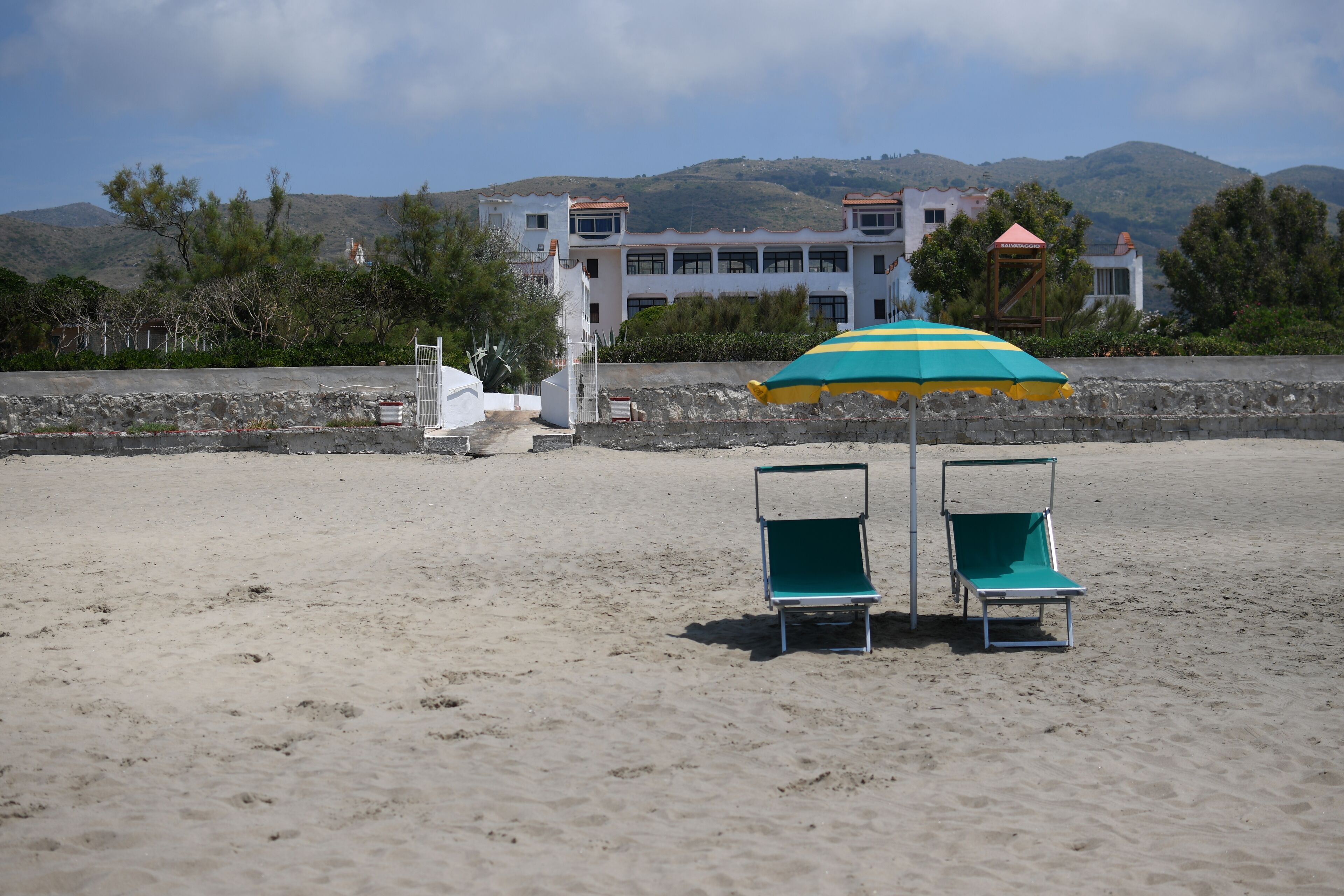 Una playa con dos sillas de jardín verdes y un paraguas a rayas, un edificio blanco al fondo y un paisaje montañoso.
