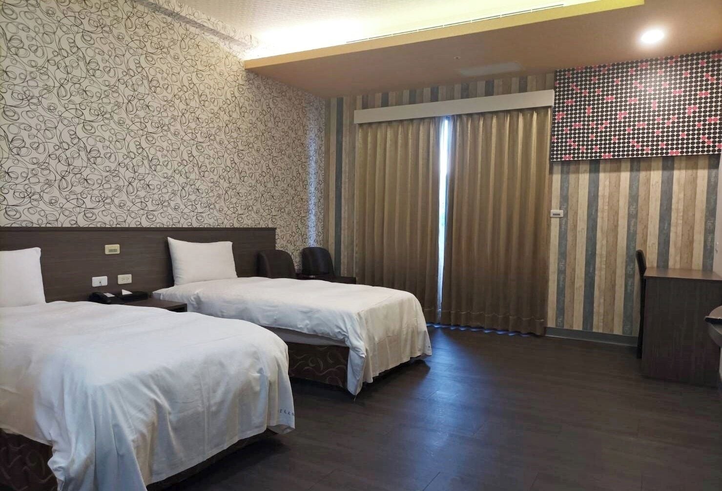 Una habitación de hotel con dos camas, un escritorio, una silla y un ventanal con cortinas.