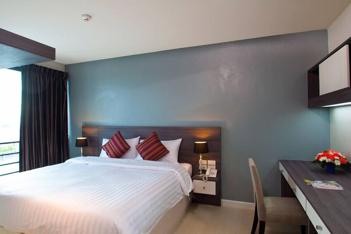 Habitación de hotel con una cama grande, un escritorio y una silla. Hay una ventana con cortinas, una mesita de noche con una lámpara y un jarrón con flores en el escritorio.