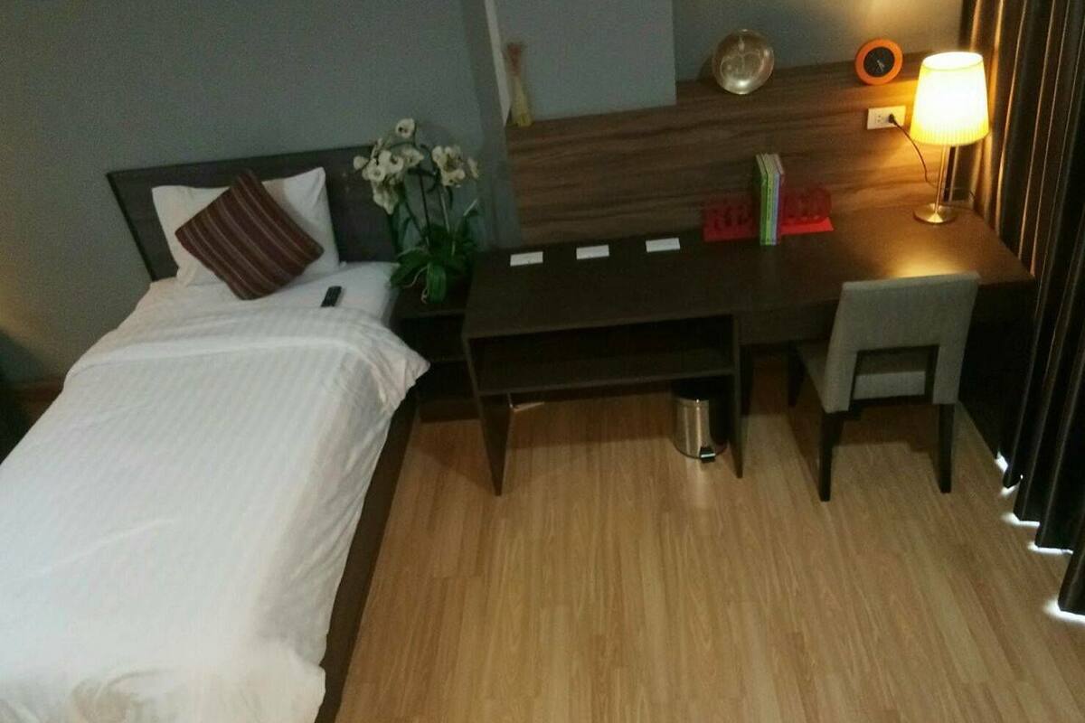 Habitación de hotel con cama, escritorio, silla, lámpara y mesita de noche con un florero.