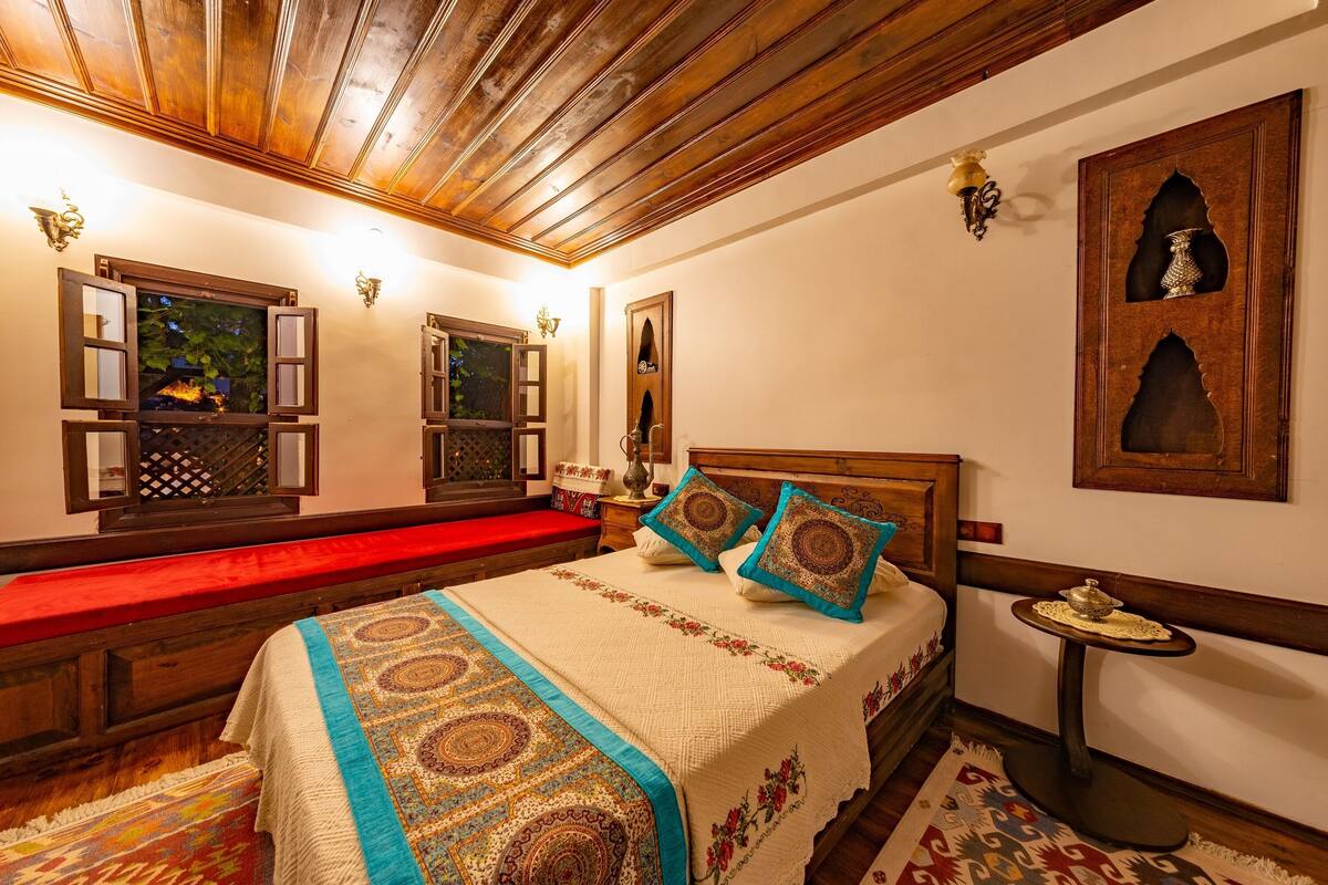 Una habitación tradicional con techo de madera, cama con ropa de cama estampada, un banco rojo y apliques decorativos en la pared.