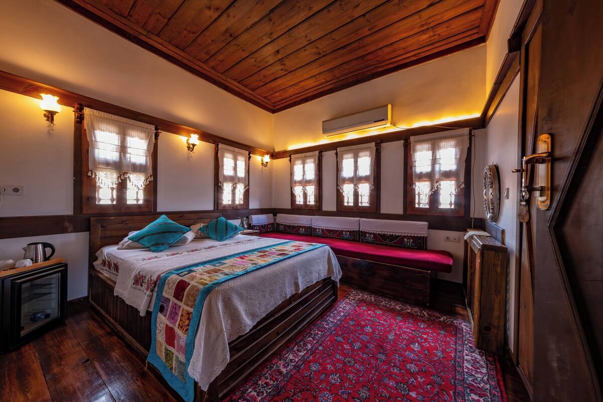 Una habitación tradicional con techo de madera, una cama grande con colcha, un banco y una alfombra roja.