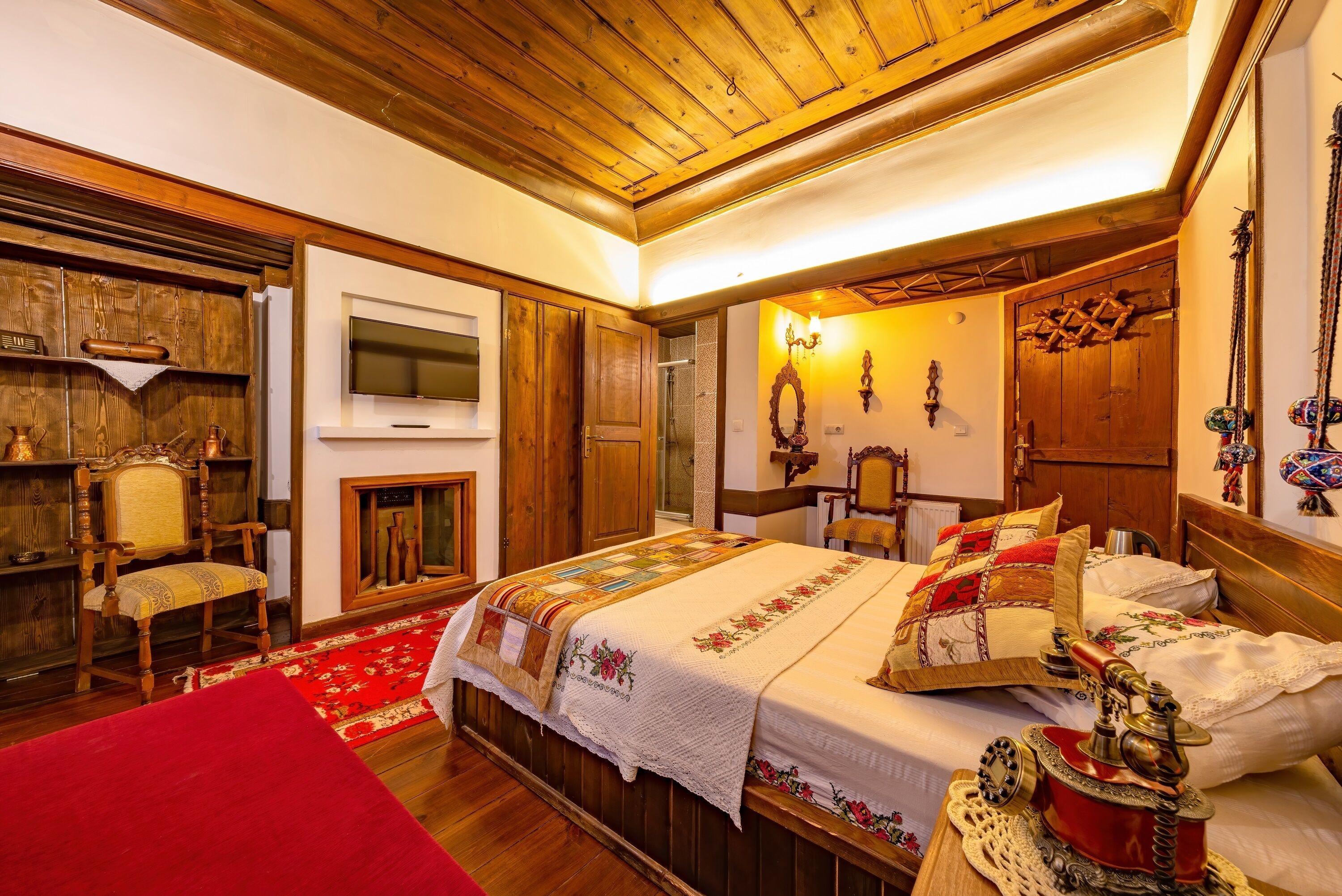A traditional room with a wooden ceiling, a fireplace, a bed with multiple pillows and a quilt, a wooden chair, and a television mounted on the wall.