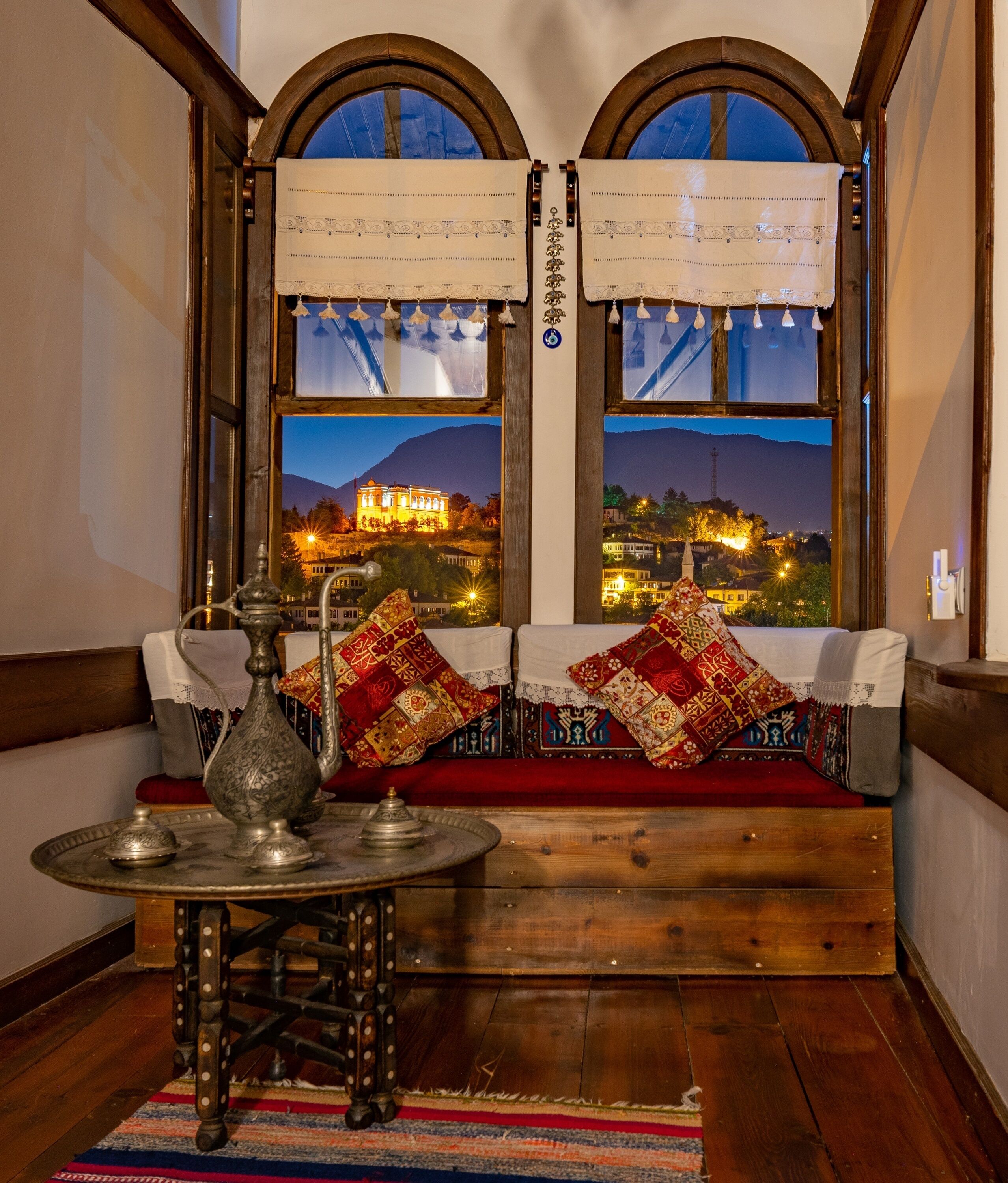 A traditional room with a wooden bench, ornate cushions, a metal tray table, and a view of a cityscape at dusk.