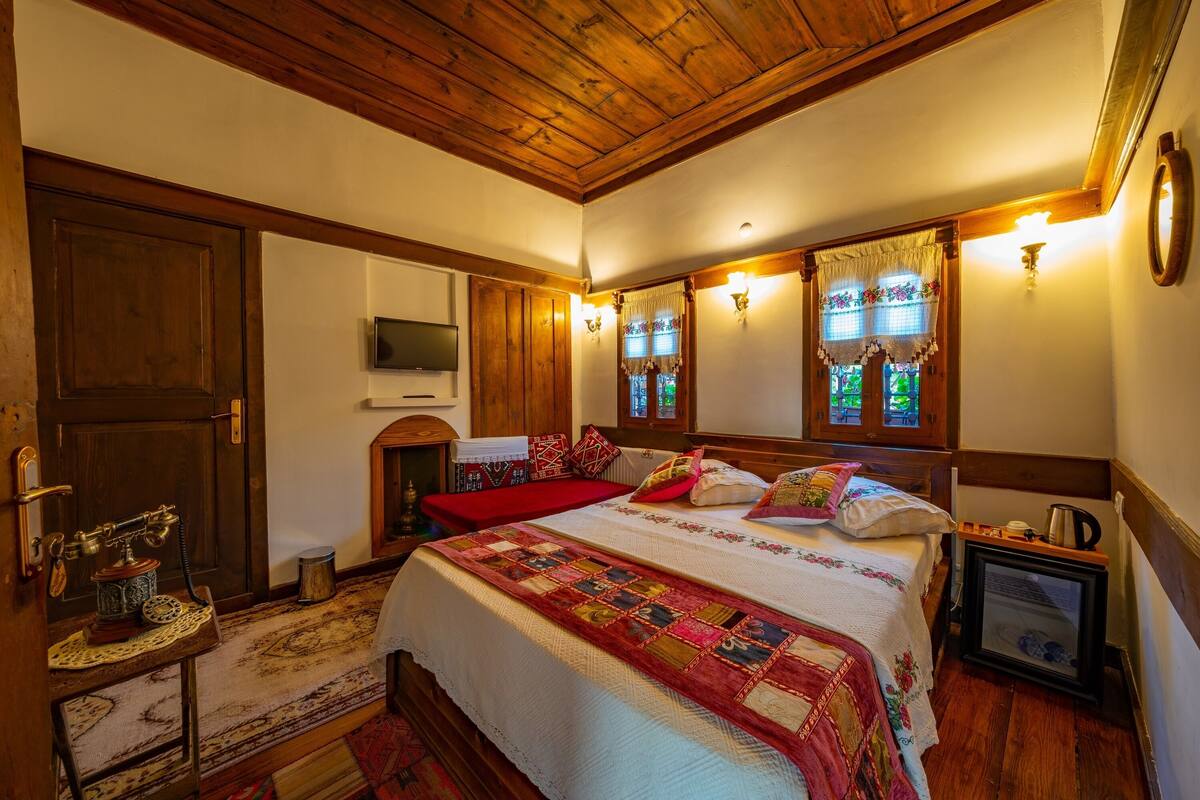 Una habitación tradicional con techo de madera, una cama grande con colcha, un televisor montado en la pared y una pequeña mesa con lámpara.