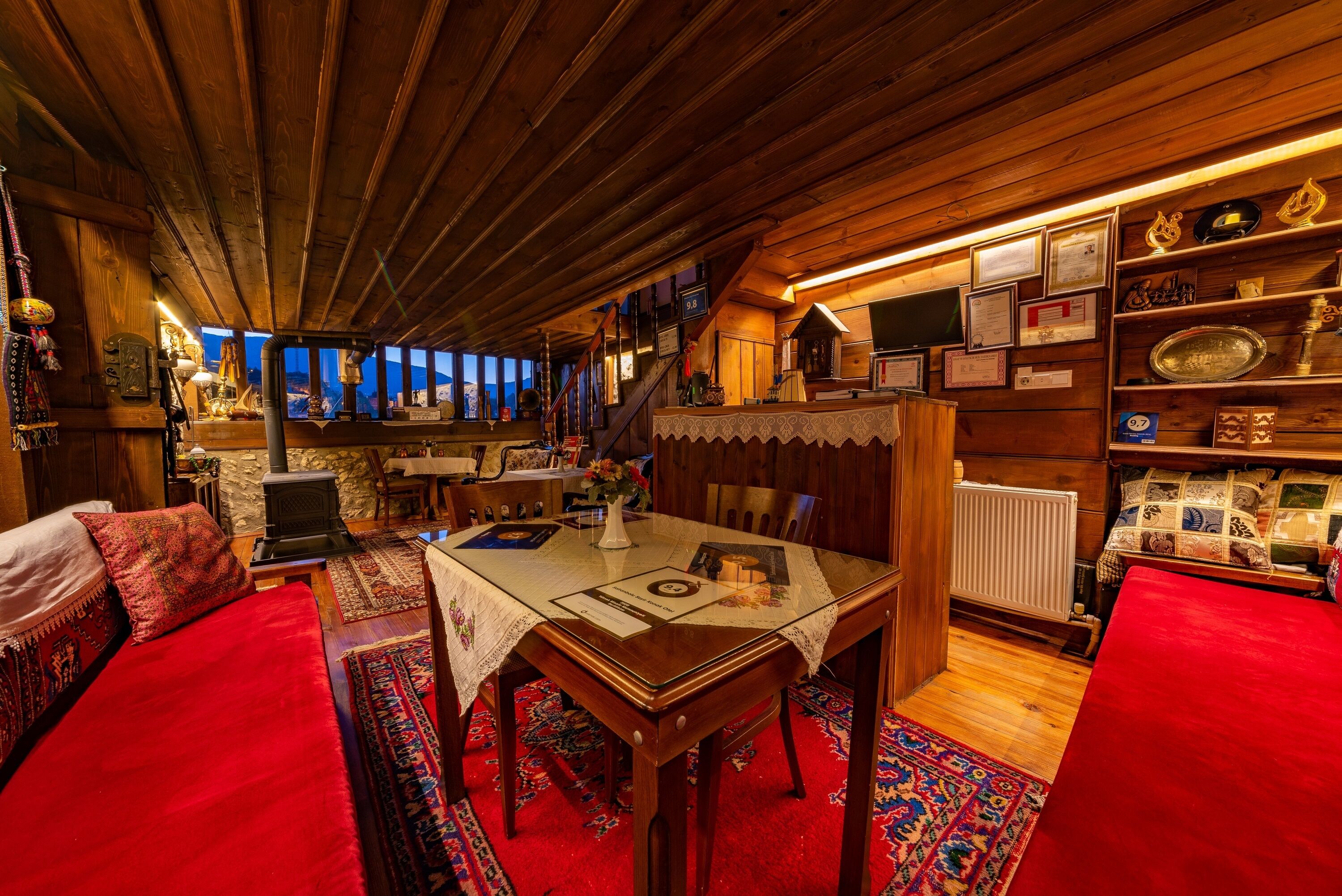 Una habitació acollidora amb sostre de fusta, una taula de dinar amb cambrera de puntes, un sofà vermell i una biblioteca amb diversos objectes.