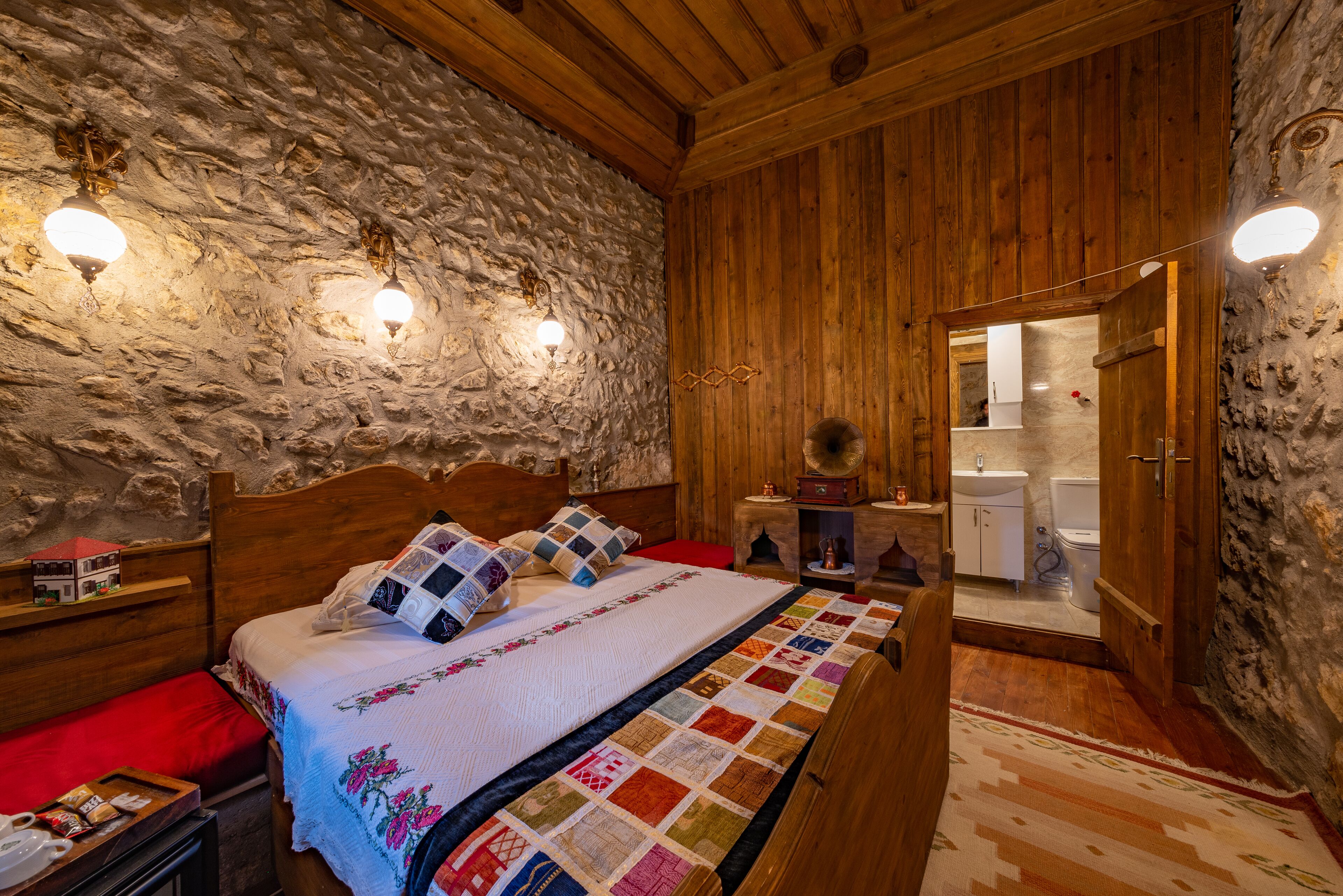 Un dormitorio con pared de piedra, techo de madera, cama con colcha, mesitas de noche y un velador.