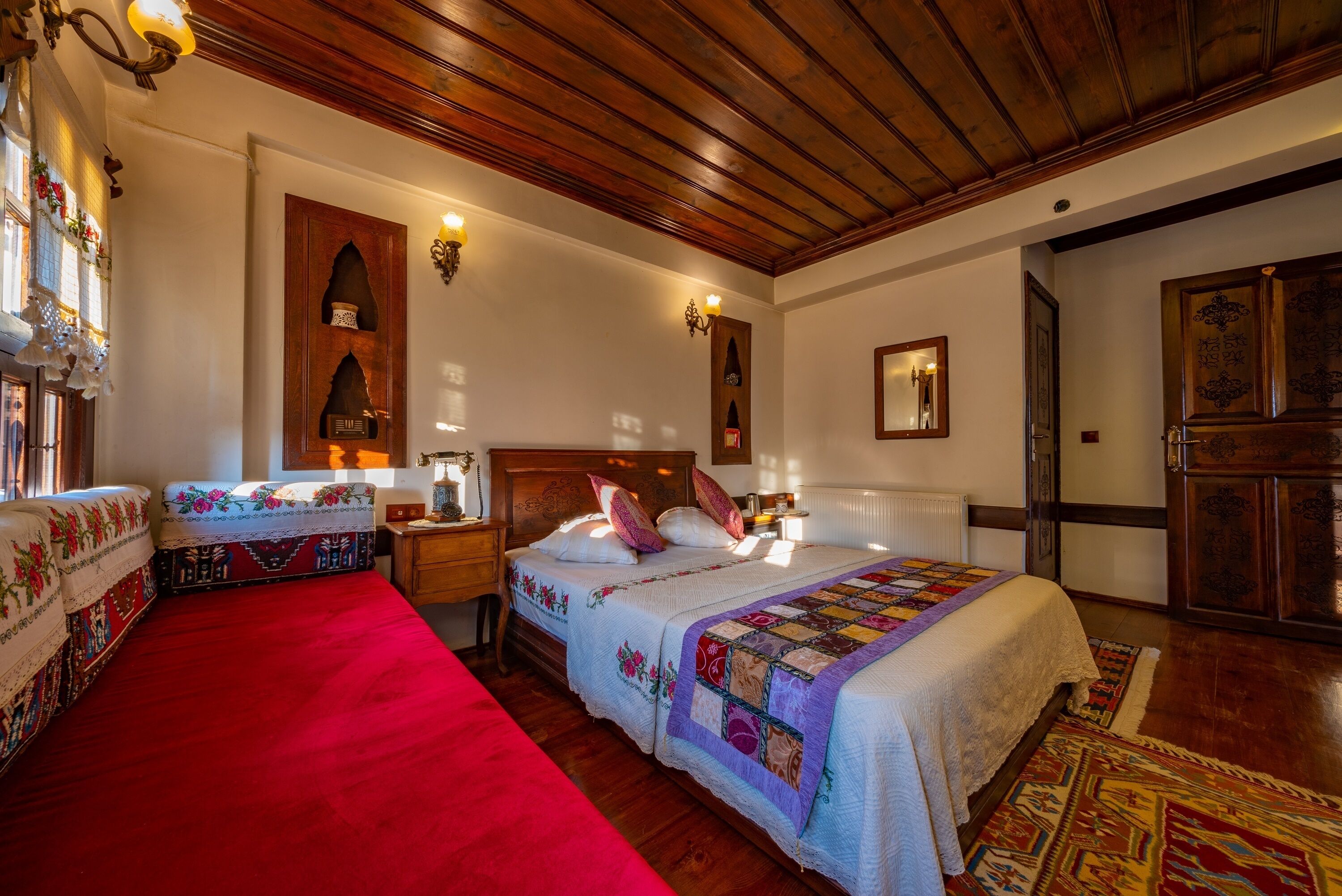 A traditional room with a wooden ceiling, a large bed with a quilt, a red carpet, and a wooden wardrobe.