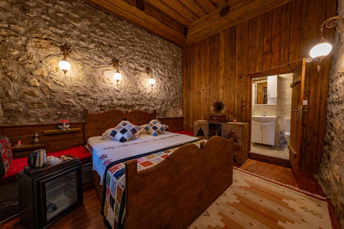 Una habitación de madera con pared de piedra, una cama con colcha, un mesita de noche, un espejo y un baño pequeño visible a través de una puerta abierta.