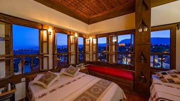 Quarto com uma cama grande, um banco vermelho e várias janelas com vista para uma cidade e montanhas.