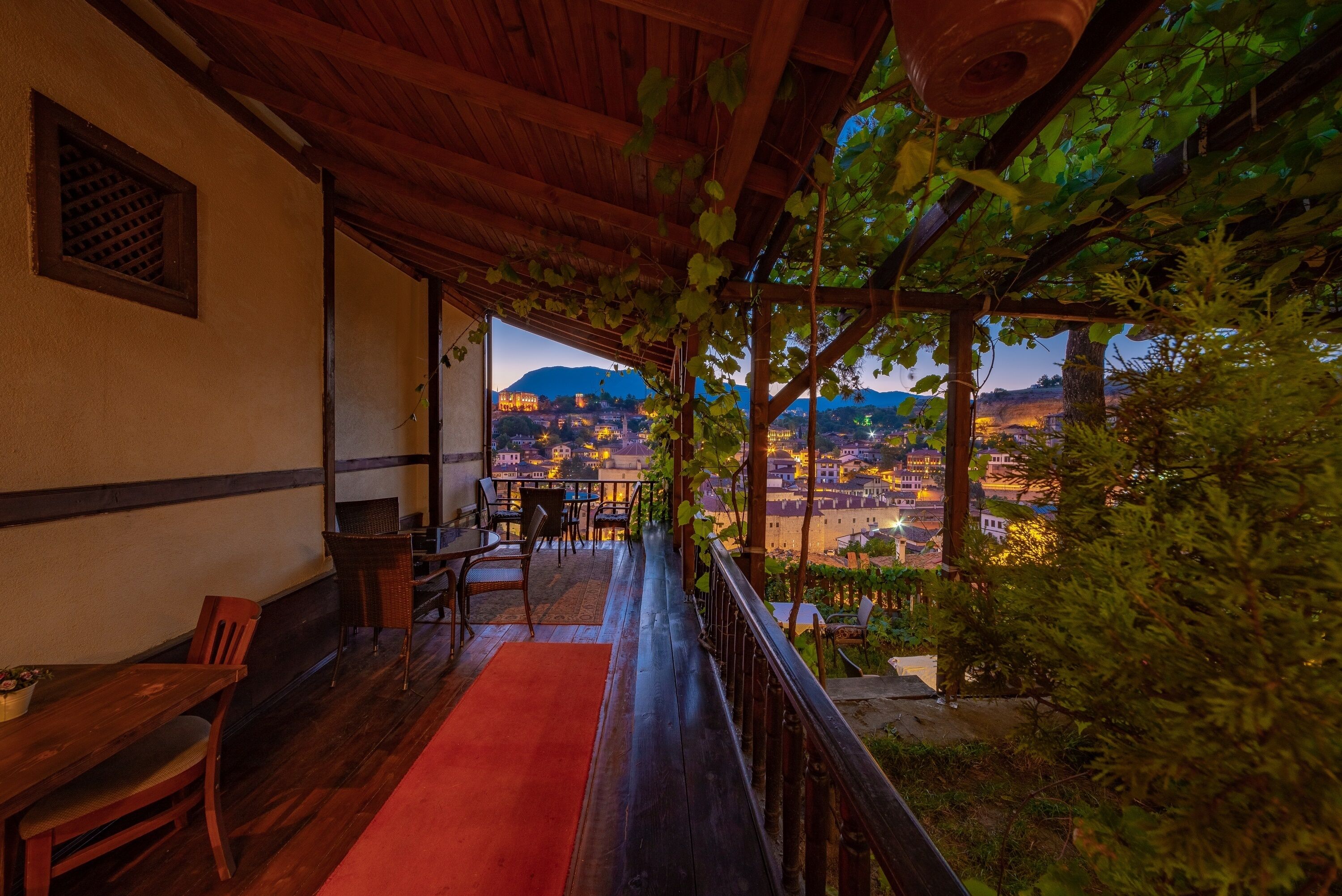 A wooden deck with a red carpet, wooden chairs, and a table, overlooking a cityscape with buildings and a mountain in the background.